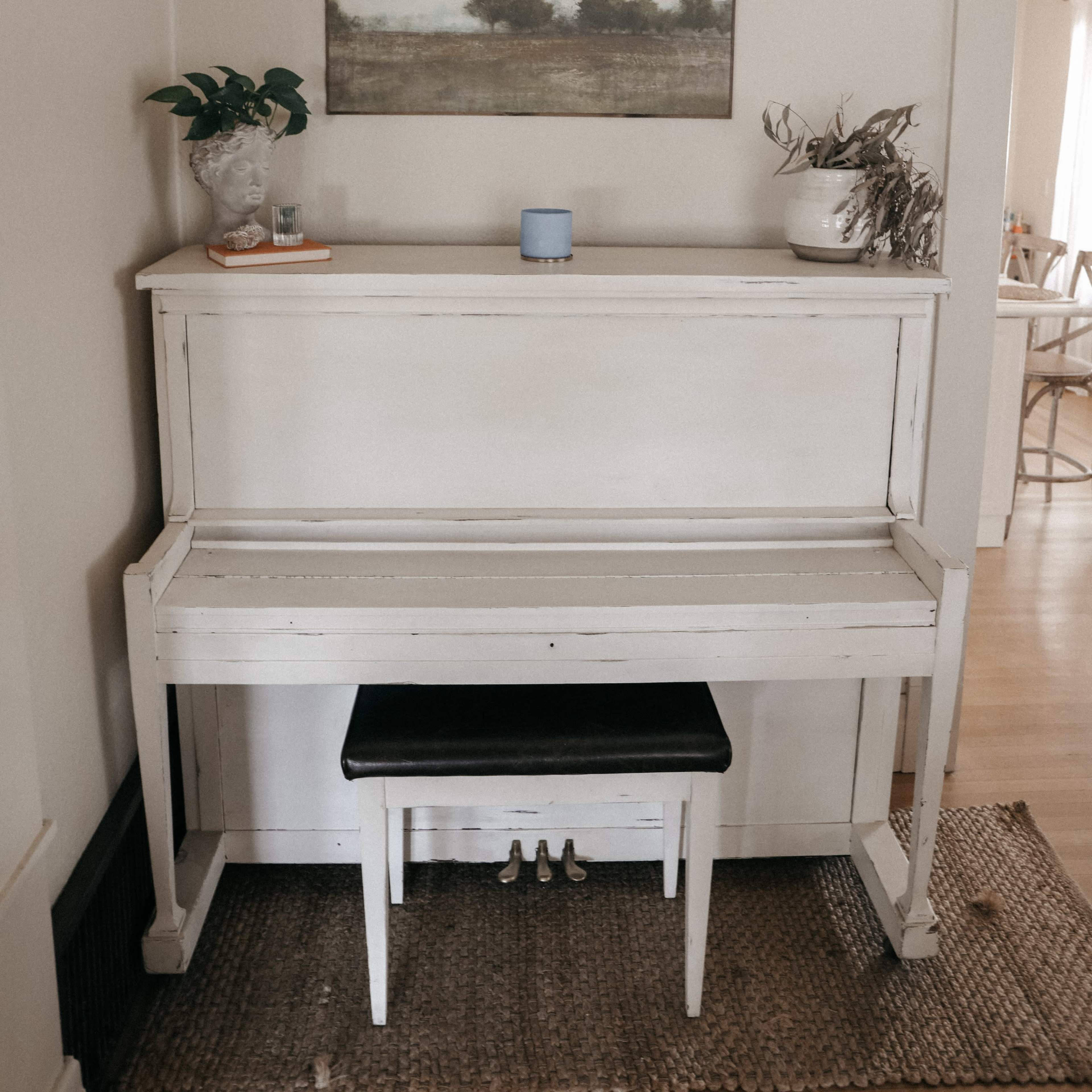 A white piano with a black bench is positioned against a light-colored wall, adorned with a painting and potted plants.