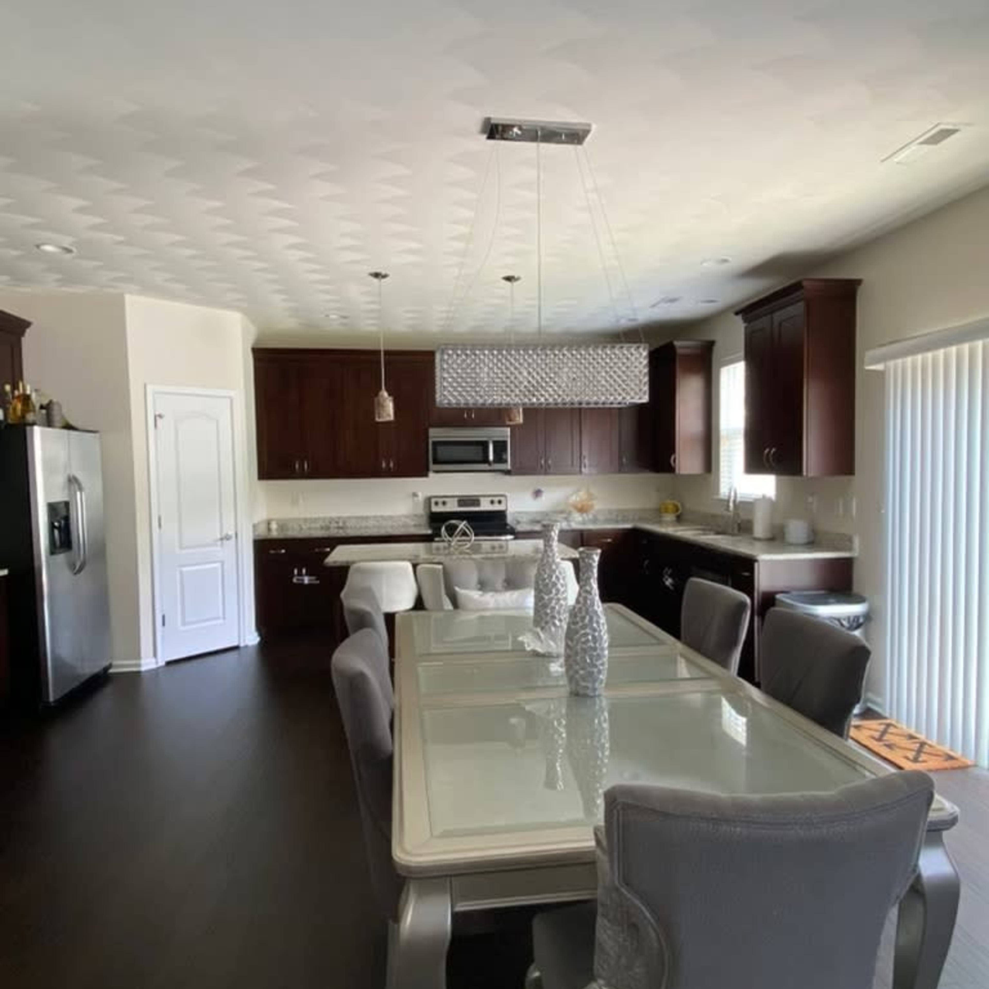 A modern kitchen and dining area featuring a rectangular glass dining table surrounded by upholstered chairs, with dark wood cabinetry and stainless steel appliances.