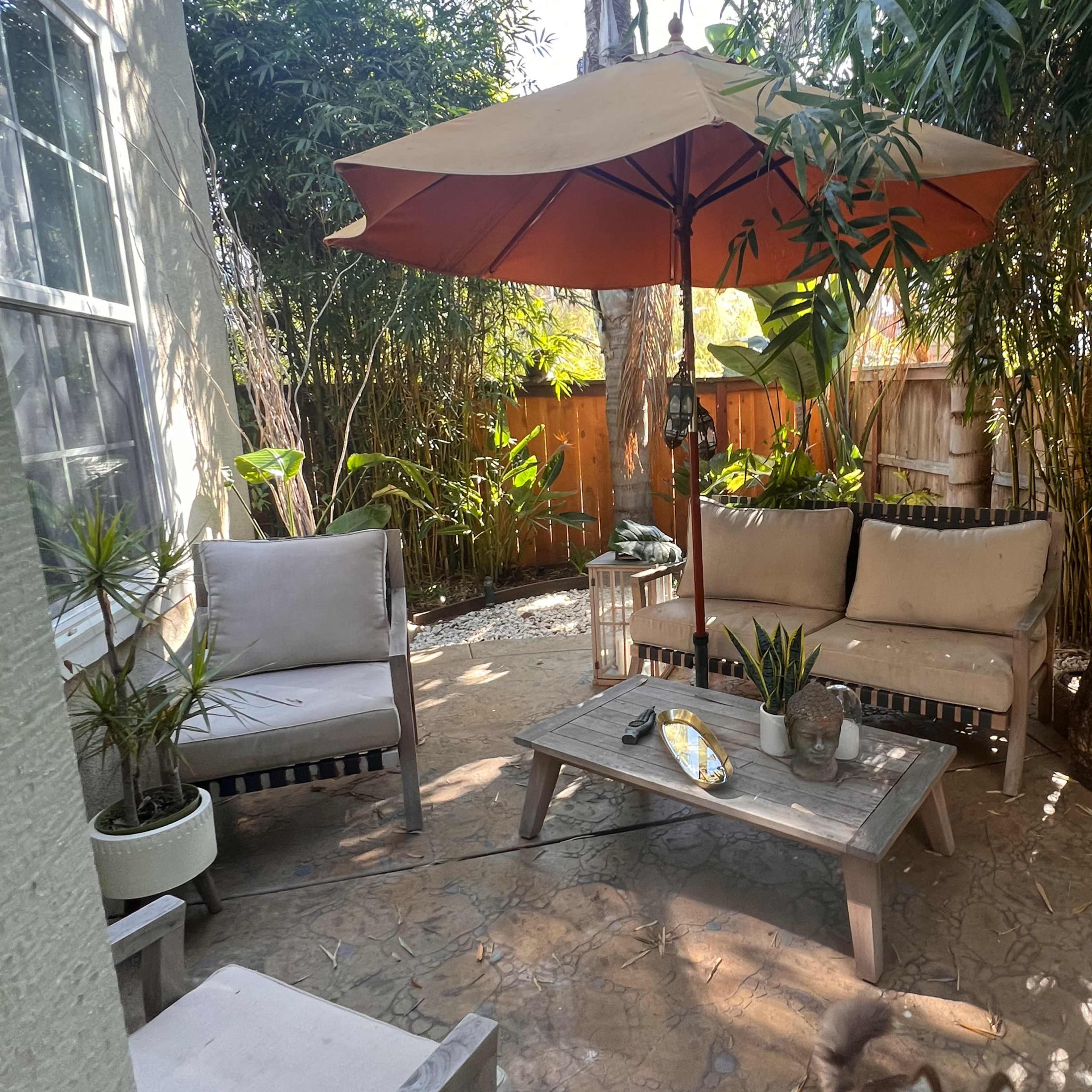 A small outdoor seating area features two chairs, a table, and an umbrella surrounded by potted plants and bamboo.