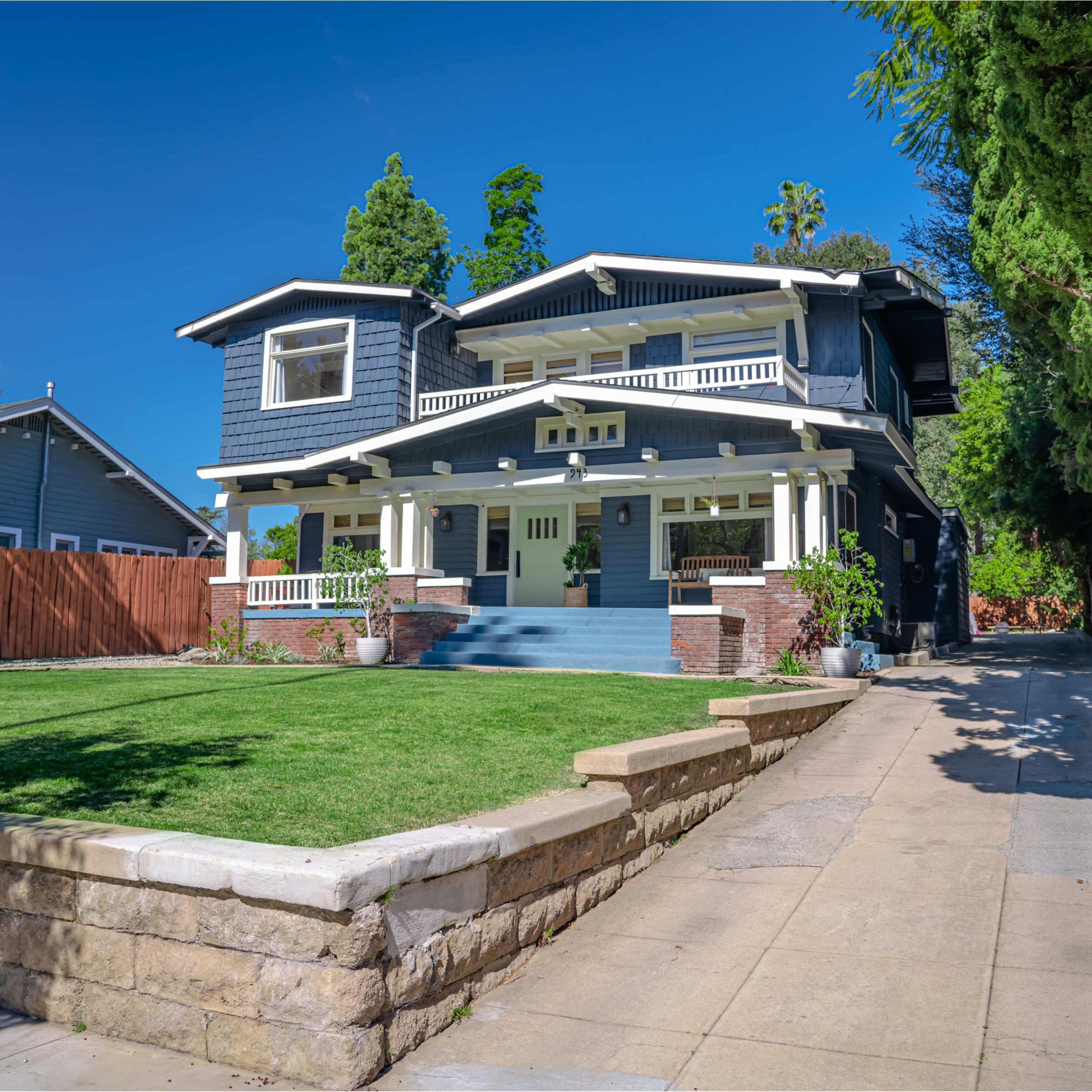 A large, two-story blue house with a welcoming front porch and neatly landscaped yard is situated on a sloped driveway.