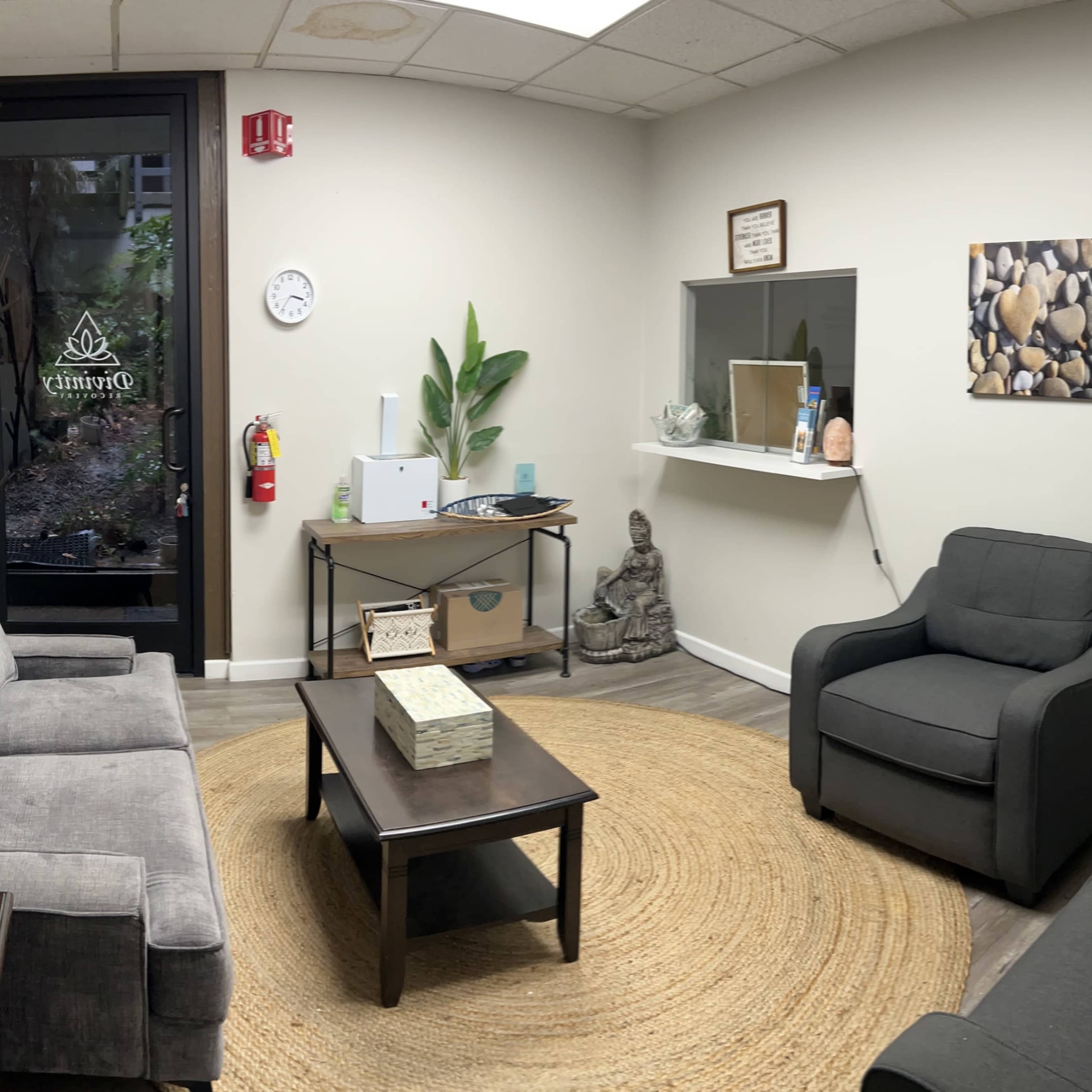 A waiting room with gray sofas, a round rug, a coffee table, and decorative items, including a small statue and plants, alongside a glass door leading outside.