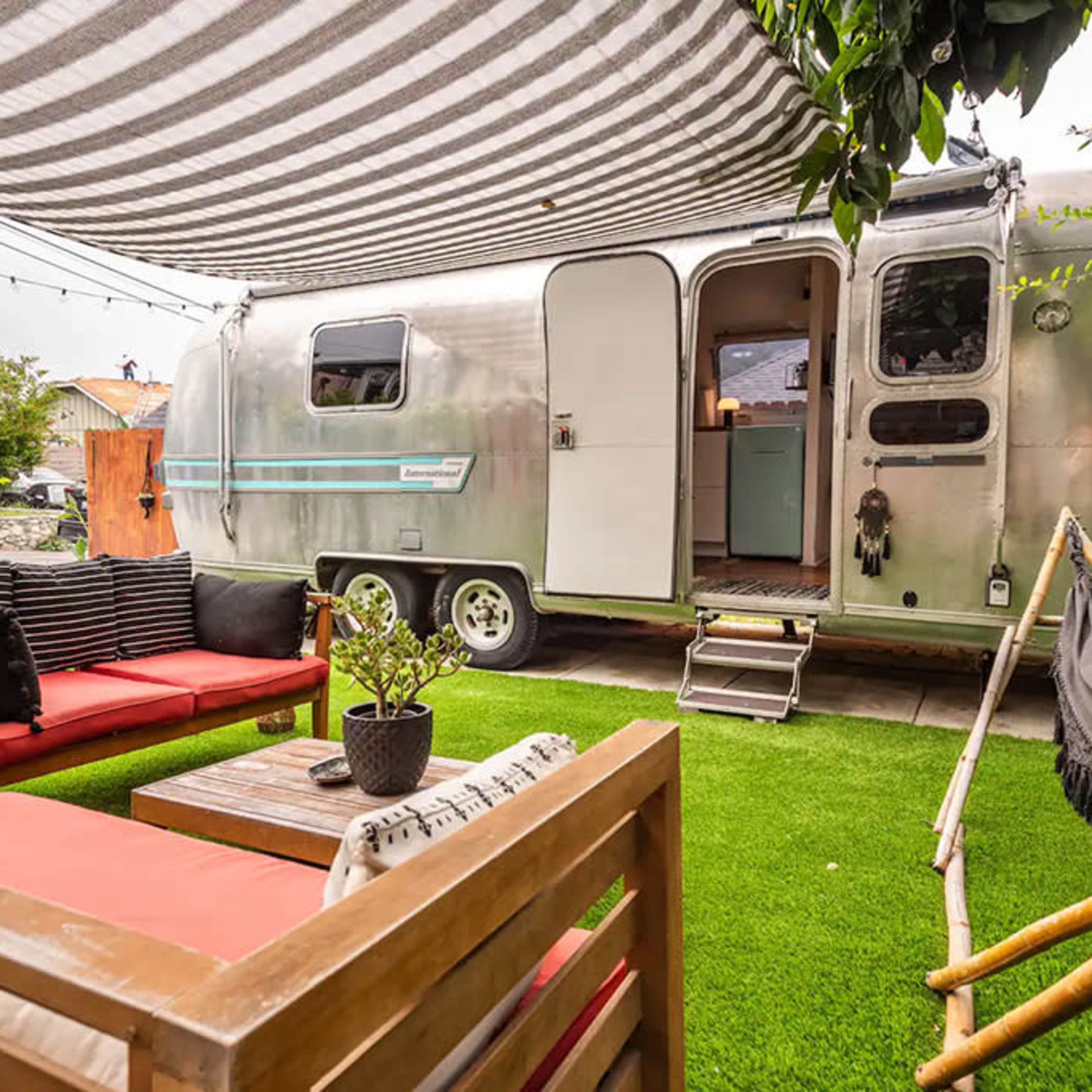 A silver Airstream trailer is parked next to a cozy outdoor seating area with red cushions on a faux grass surface, surrounded by greenery and a striped canopy overhead.