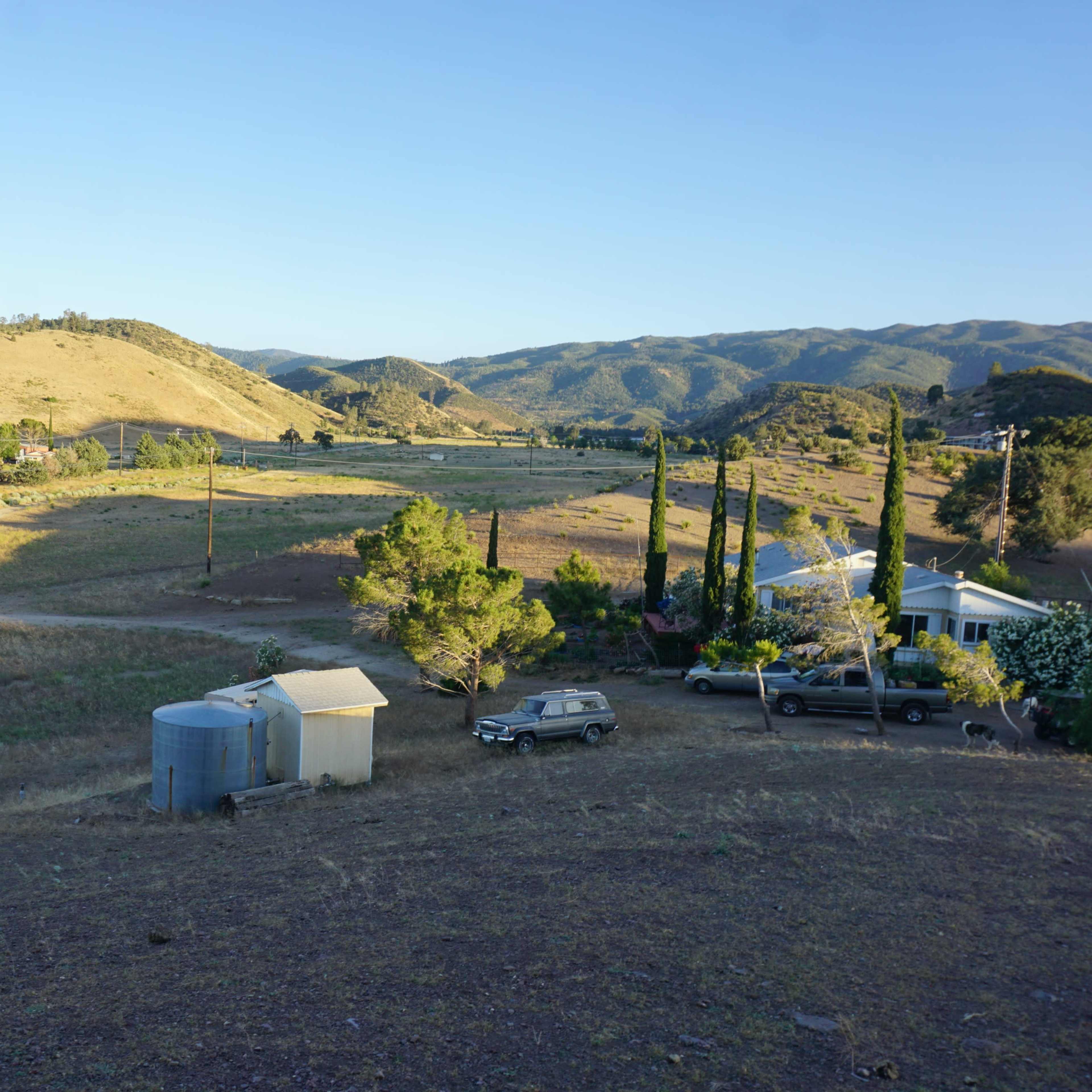 A rural landscape features rolling hills, a few scattered trees, a storage tank, and a parked vehicle near a house.