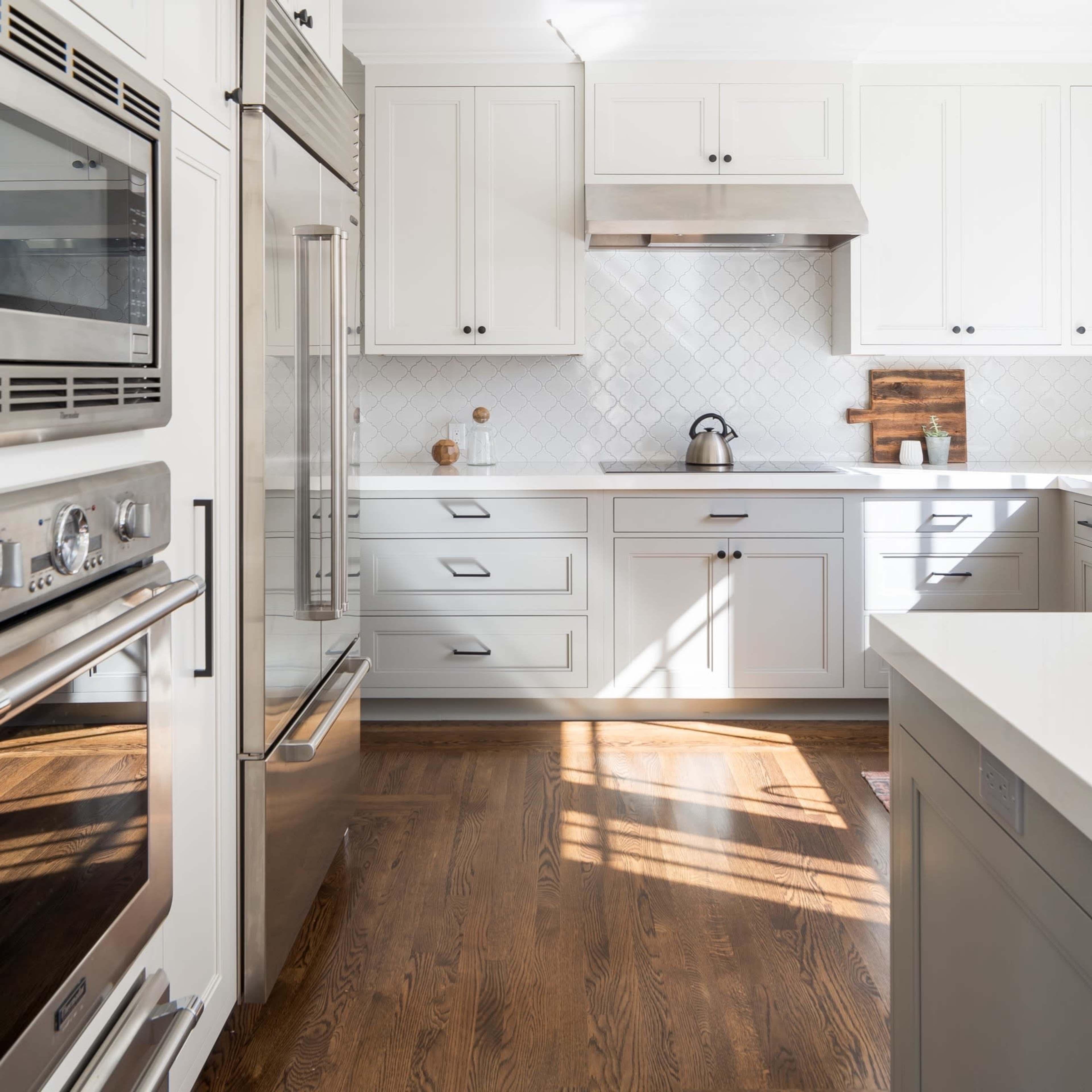 A modern kitchen featuring white cabinets, stainless steel appliances, and a hardwood floor with sunlight streaming in through a window.