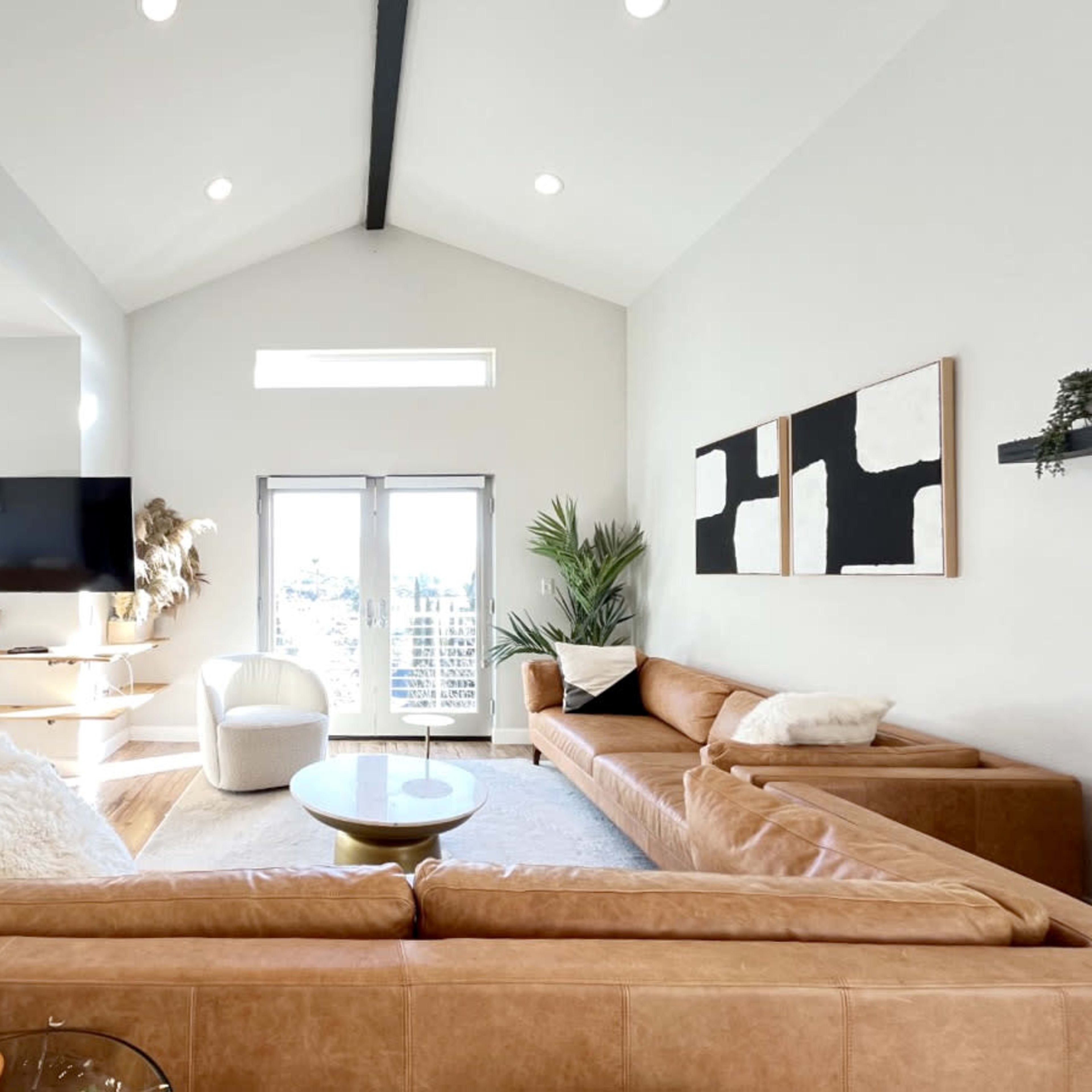 A modern living room featuring a large, brown sectional sofa, a round coffee table, a wall-mounted TV, and large windows allowing natural light to enter.