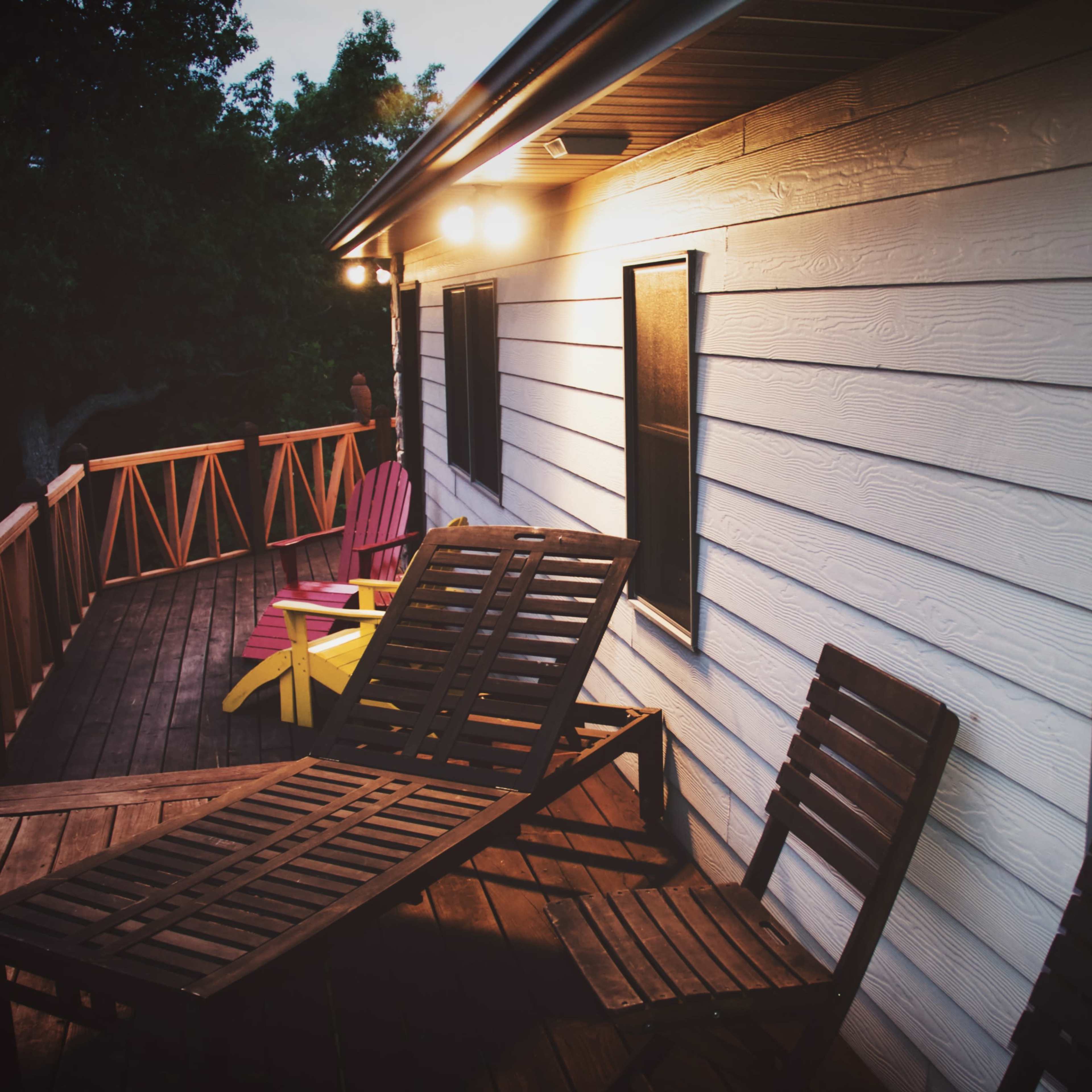 A wooden deck with two lounge chairs, colorful outdoor seating, and a house illuminated by overhead lights at dusk.