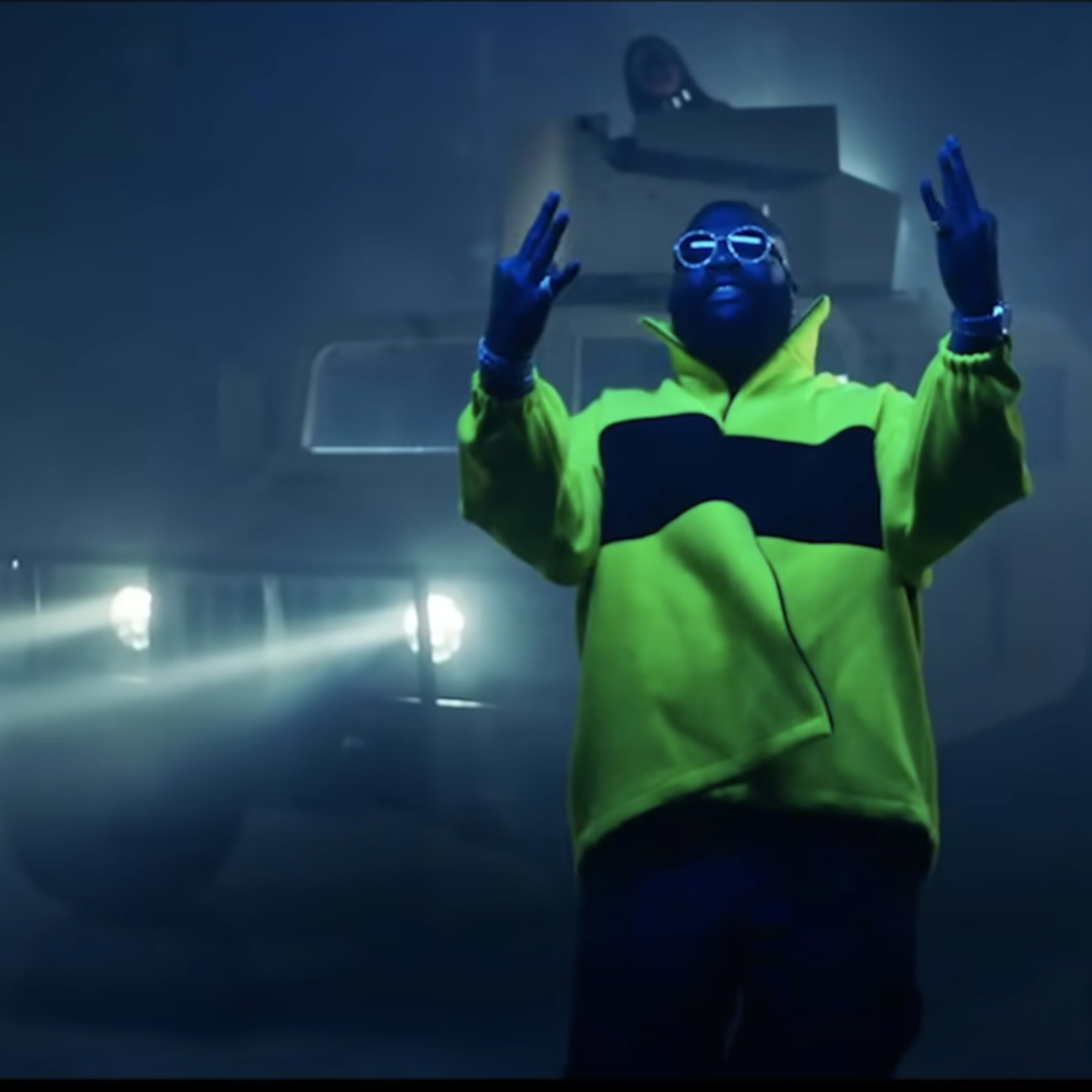 A person in a bright yellow jacket stands with arms raised in front of an illuminated vehicle in a foggy setting, while another figure is partially visible in the background.