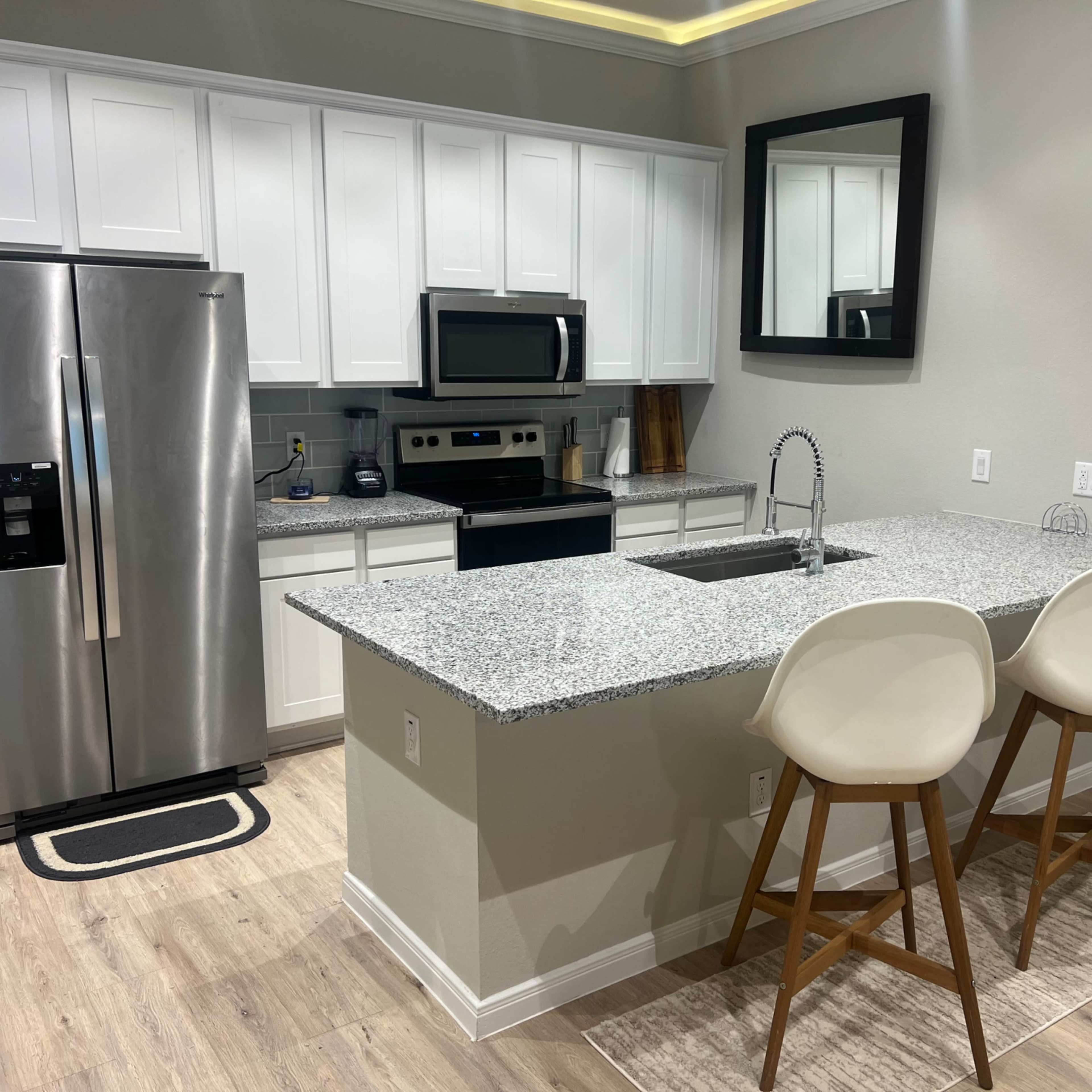 A modern kitchen features white cabinets, stainless steel appliances, a granite countertop island with seating, and a large mirror on the wall.