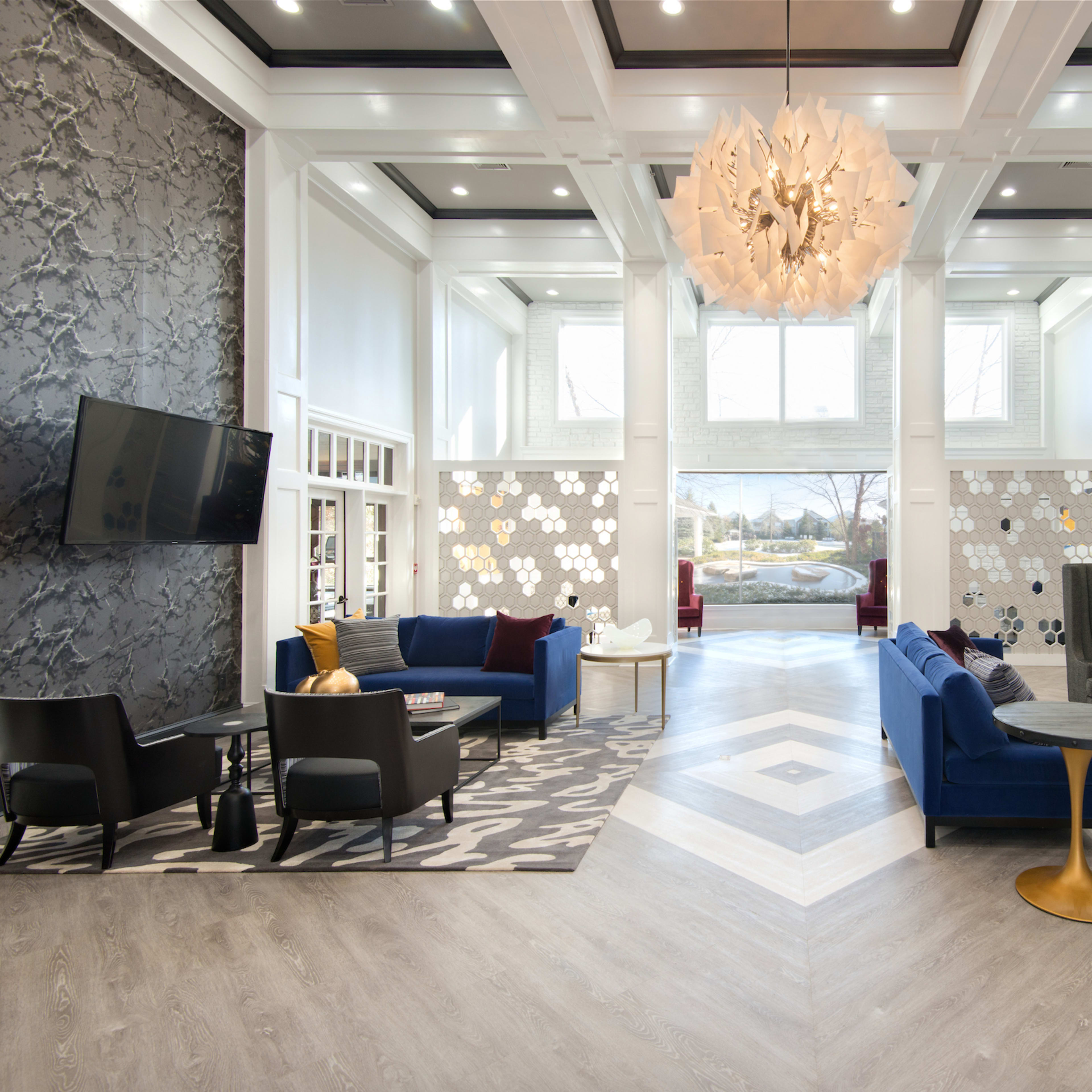 A modern lobby features seating areas with blue and black couches, a large chandelier, and a fireplace, complemented by artistic wall decor and natural light from large windows.