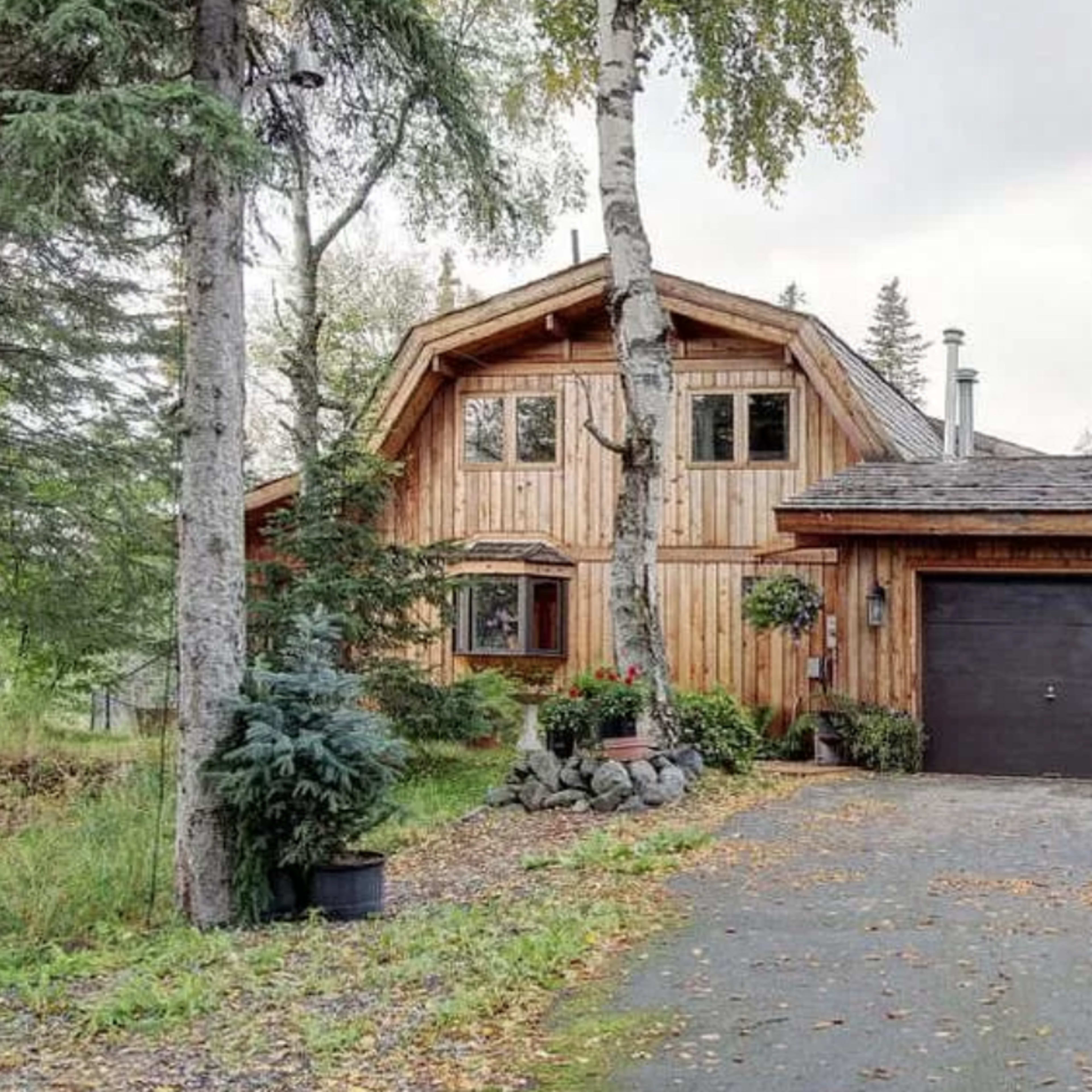 A wooden house with a steep roof and a double garage is surrounded by trees and a gravel driveway.