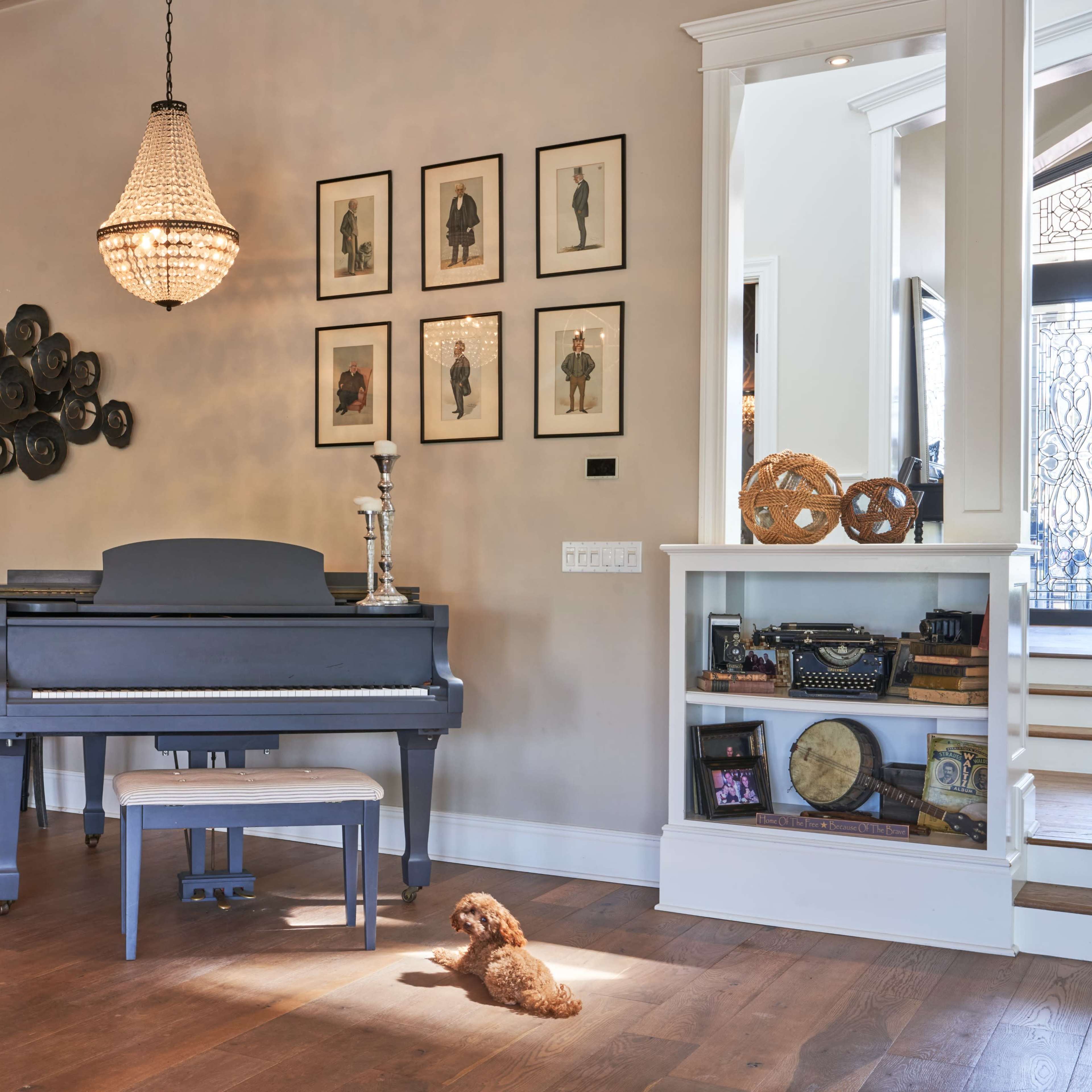 A grand piano sits in a well-lit room with framed artwork on the walls and a small dog resting on the floor nearby.