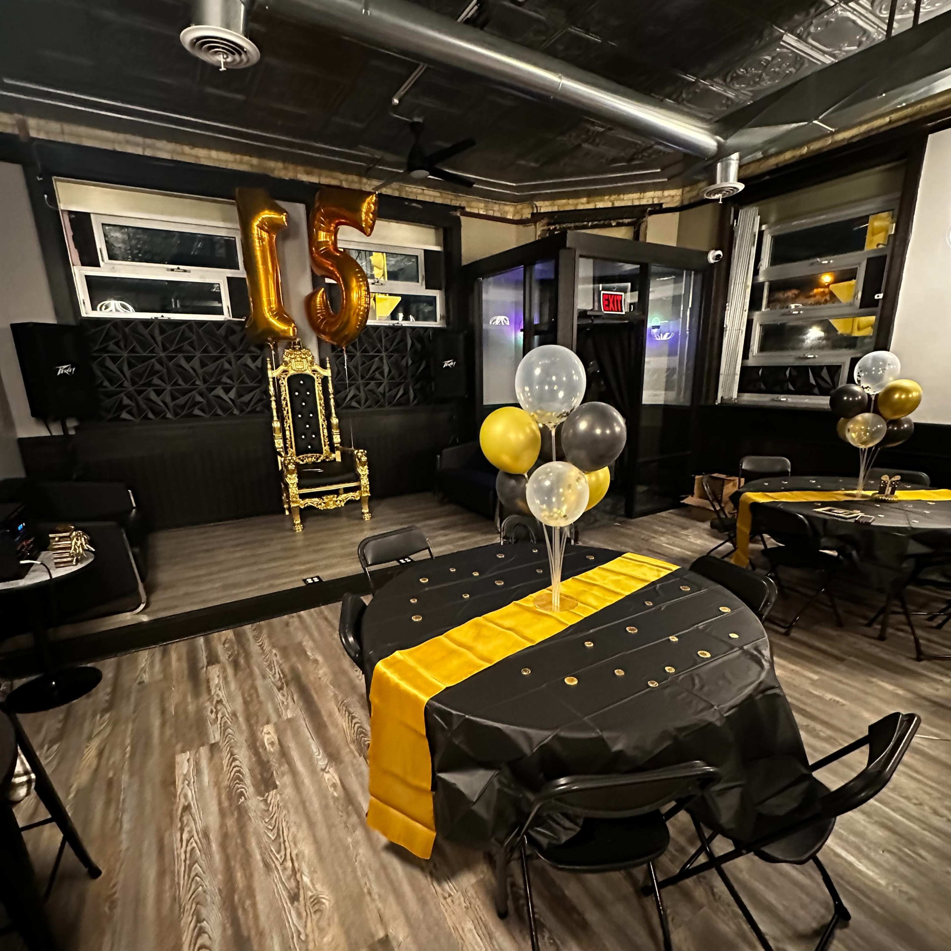 A party venue decorated with black and gold table settings, balloons, and a large "15" balloon in the background.