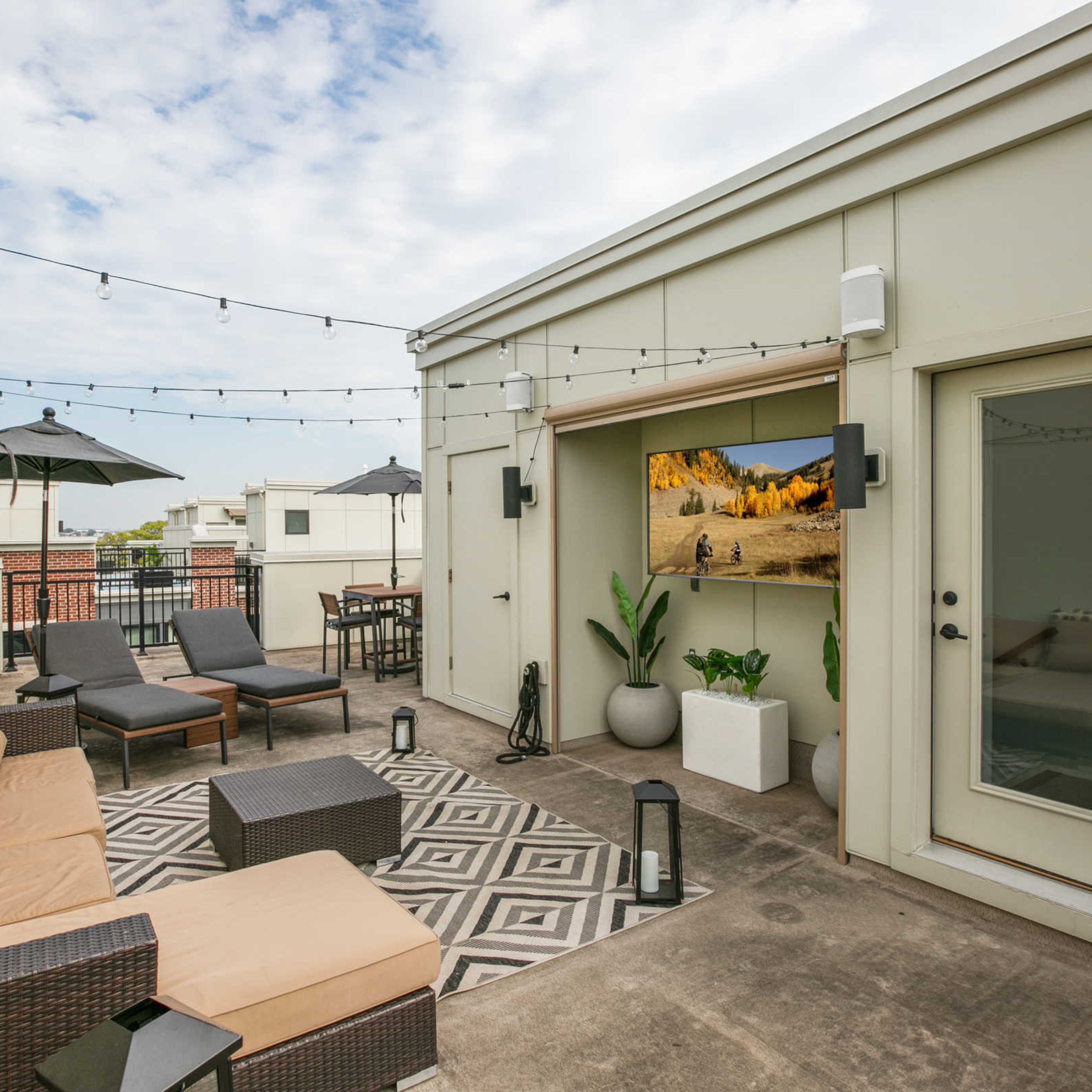 A rooftop patio furnished with a sectional sofa, lounge chairs, and decorated with string lights and potted plants.