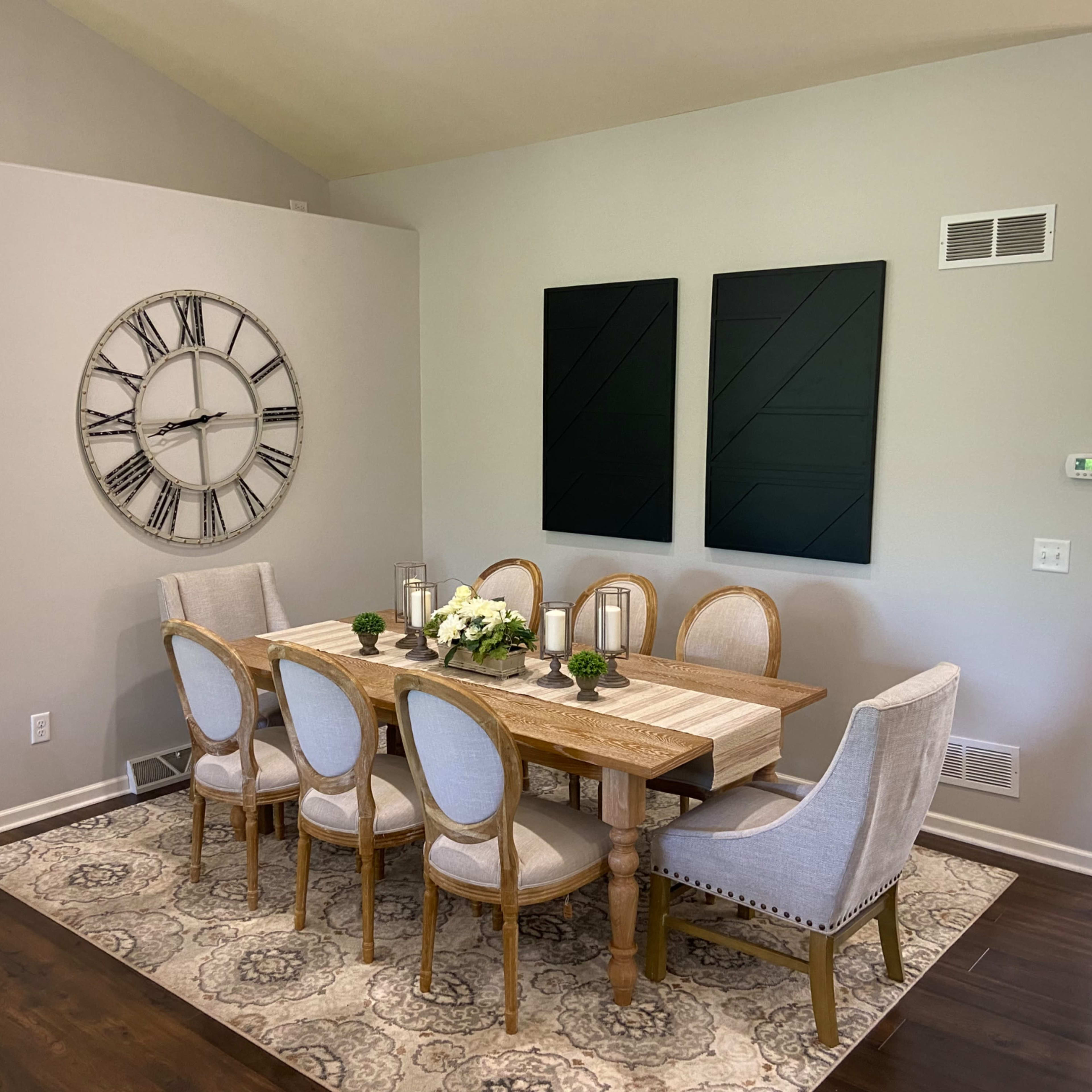 A dining room features a wooden table surrounded by eight chairs, with a large wall clock and two black wall art pieces.
