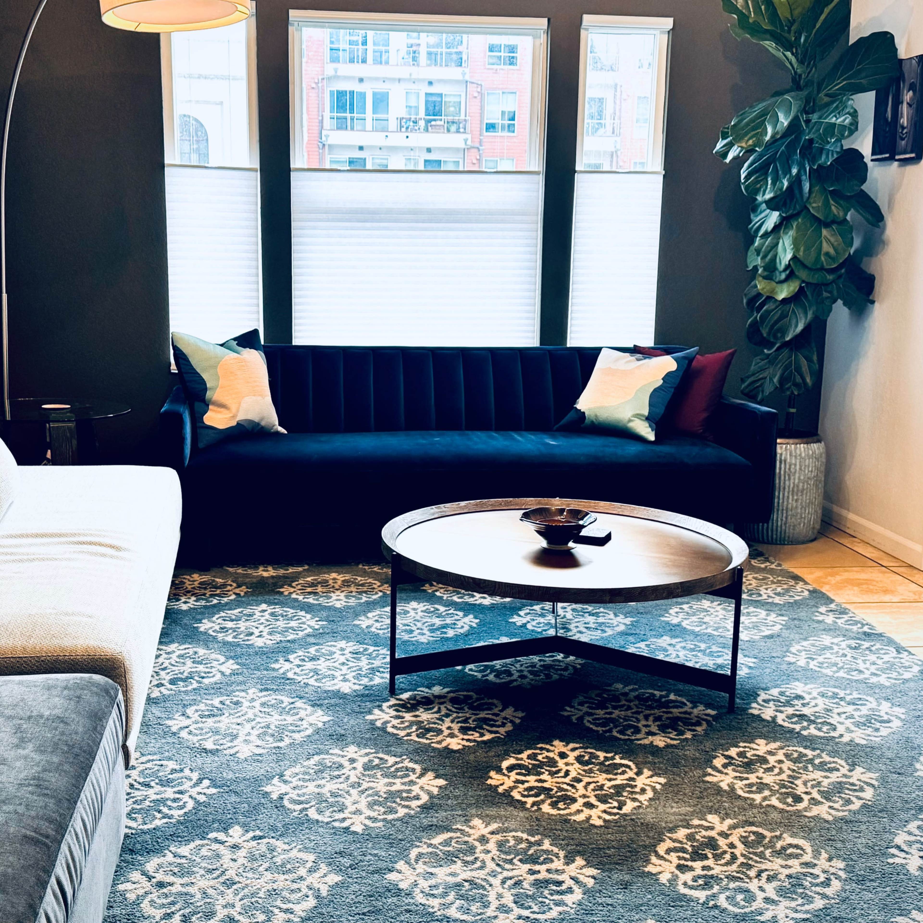 A modern living room with a light-colored sofa, a dark blue couch, a round coffee table, and large windows providing daylight illumination.