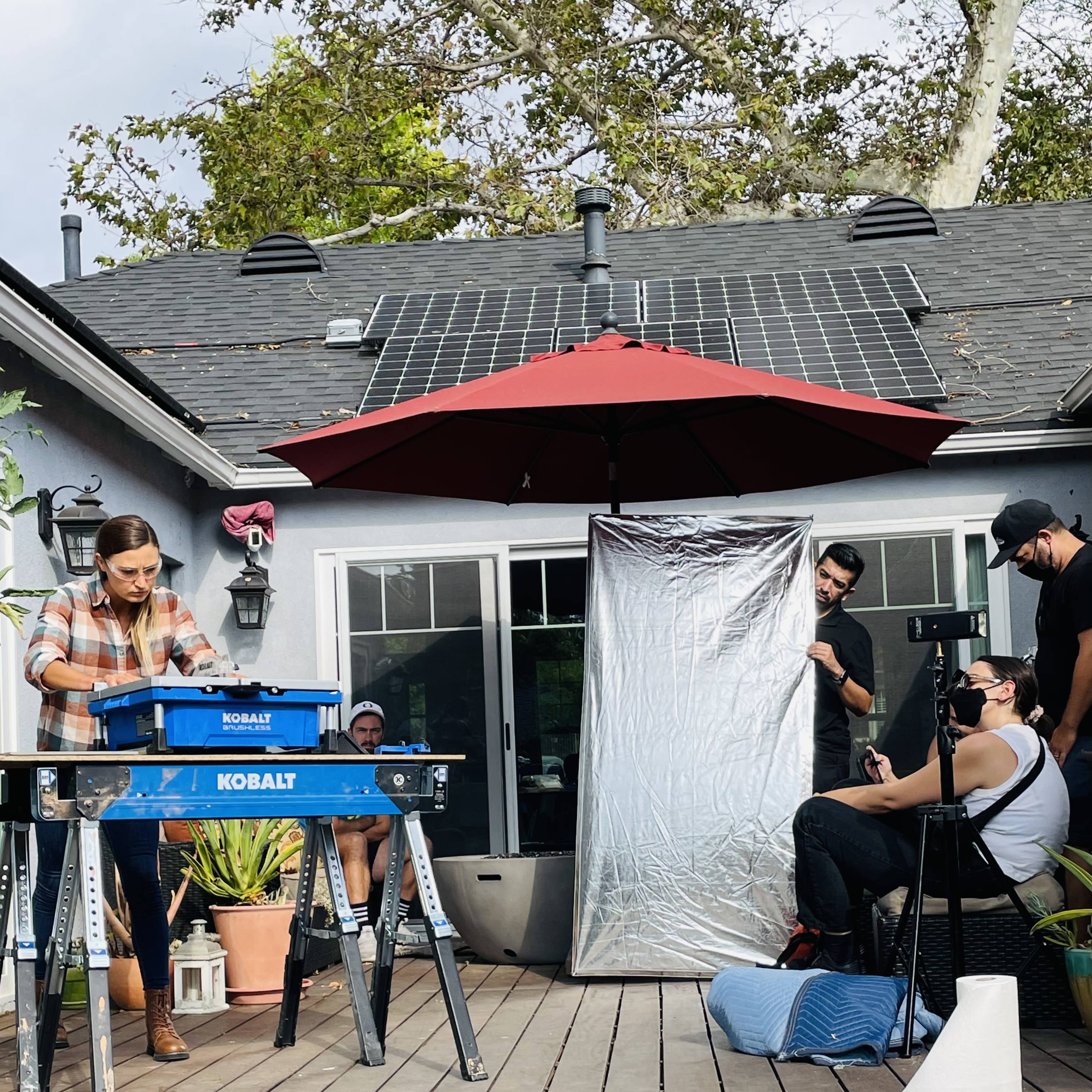 A woman works on a project at a table in a backyard while several people observe and assist.