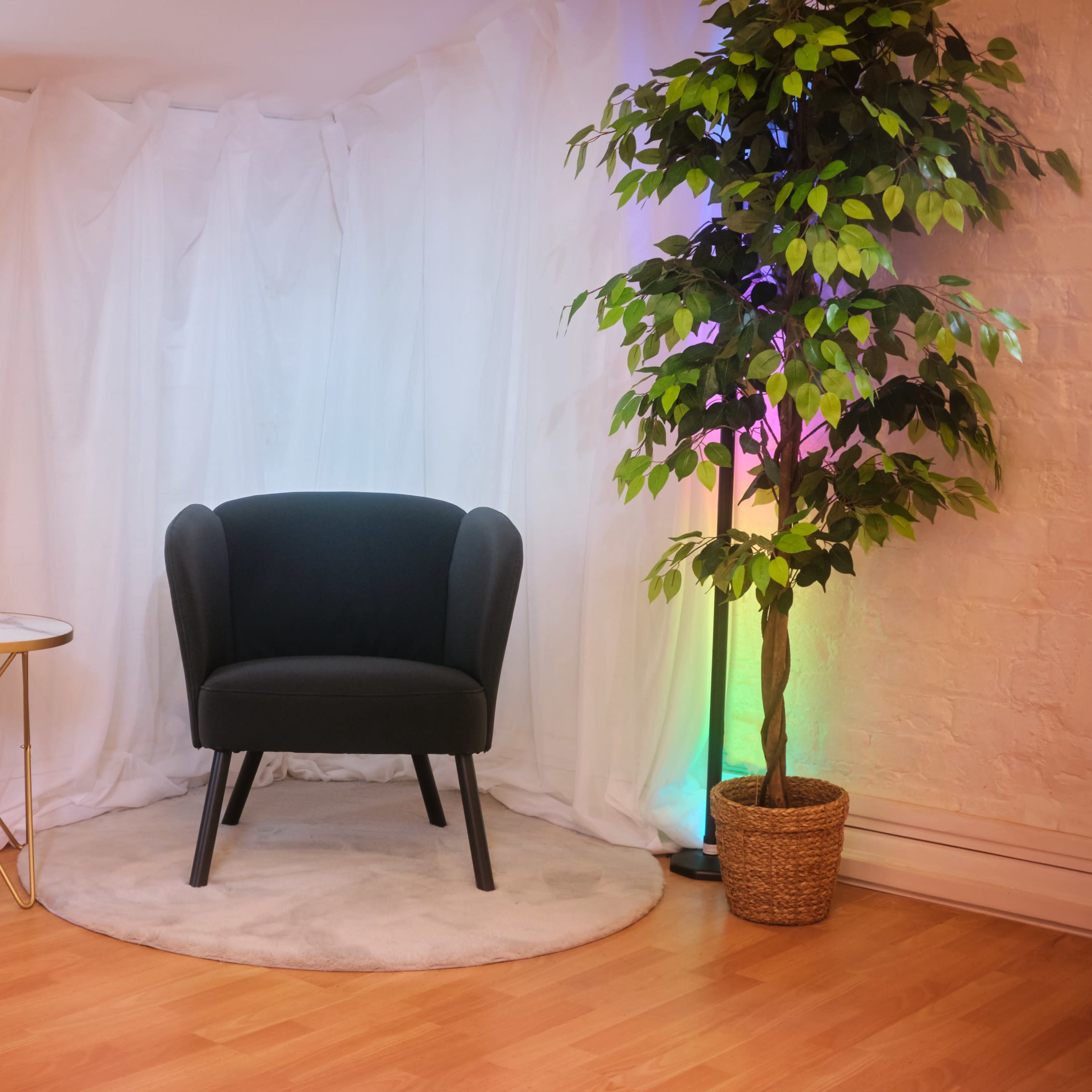 A green upholstered chair is positioned next to a round table and a large mirror, surrounded by a potted plant and draped white fabric with colorful lighting.