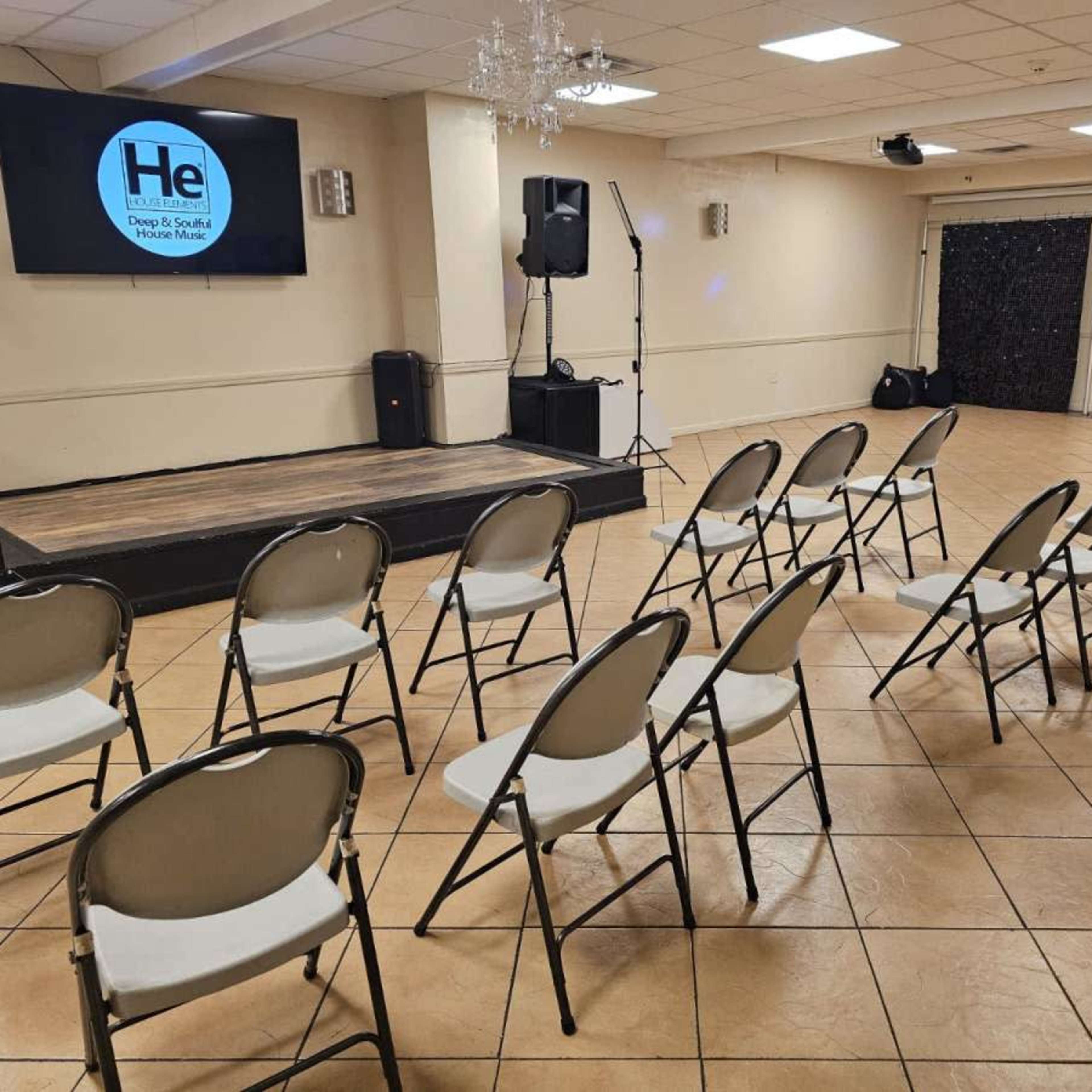 A meeting room with a raised stage, a large screen displaying a logo, and several rows of empty folding chairs arranged in front.
