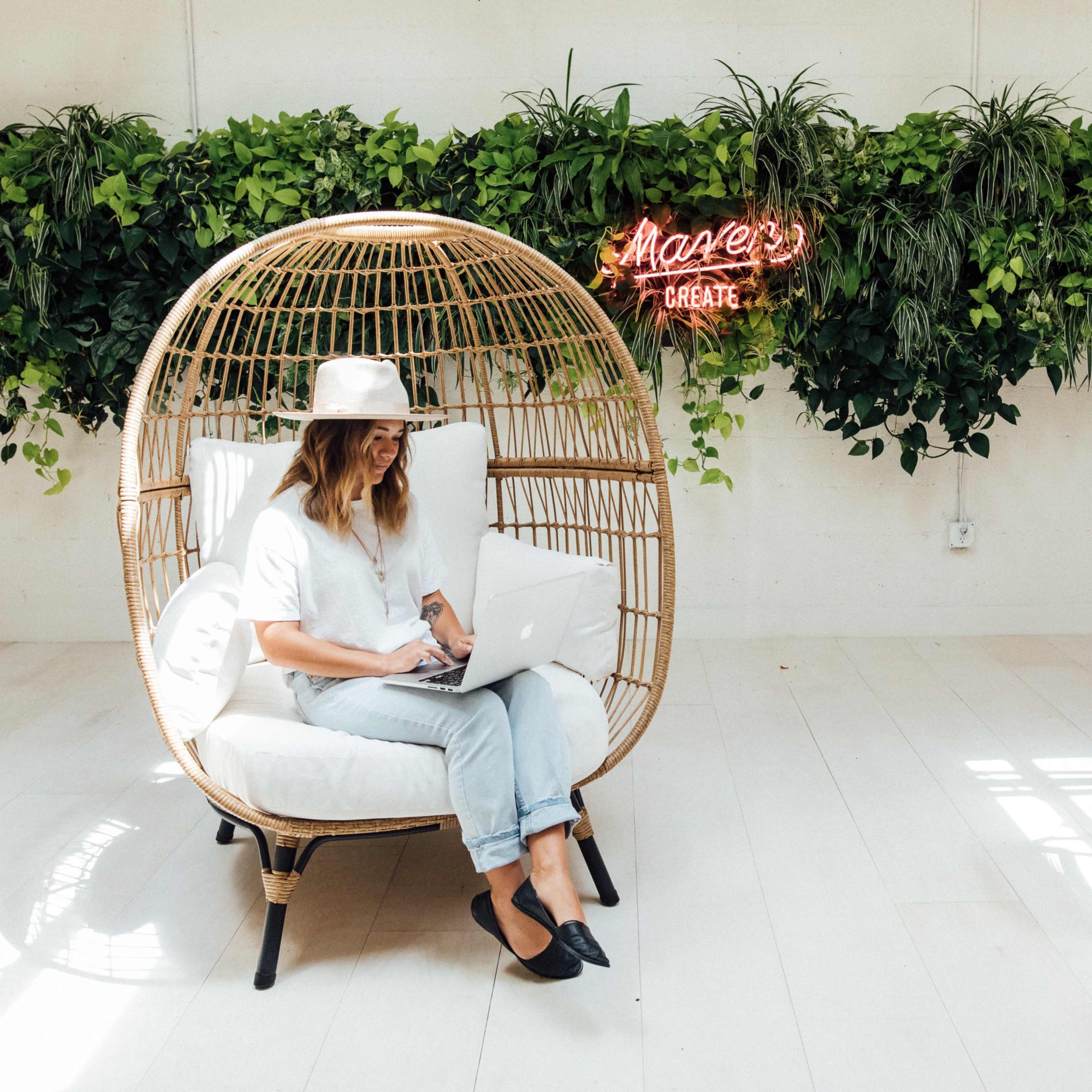 A woman sits in a large wicker chair, working on a laptop in a bright room adorned with hanging plants and a neon sign that says "Create."