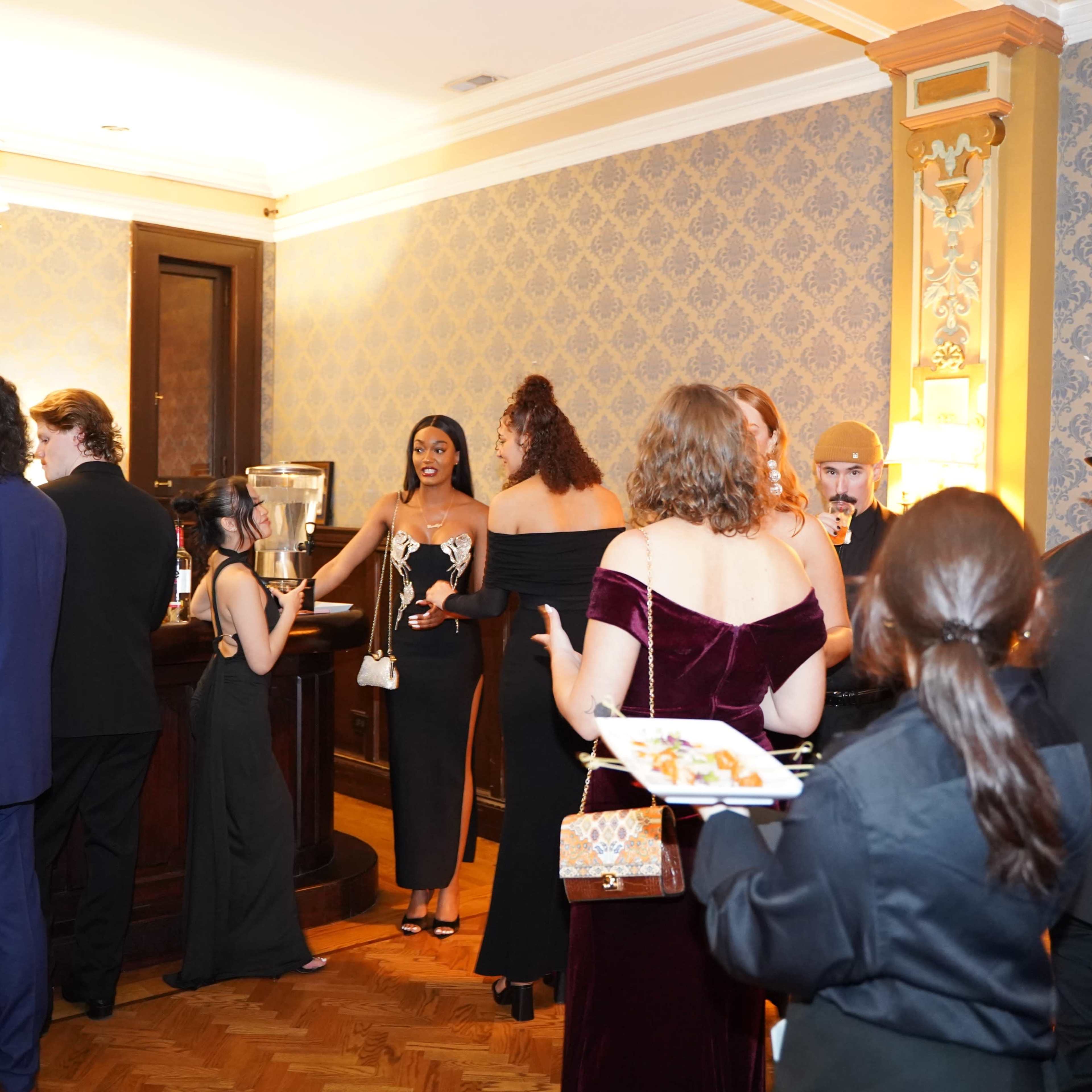 A group of elegantly dressed individuals mingle around a bar in a lavishly decorated room.