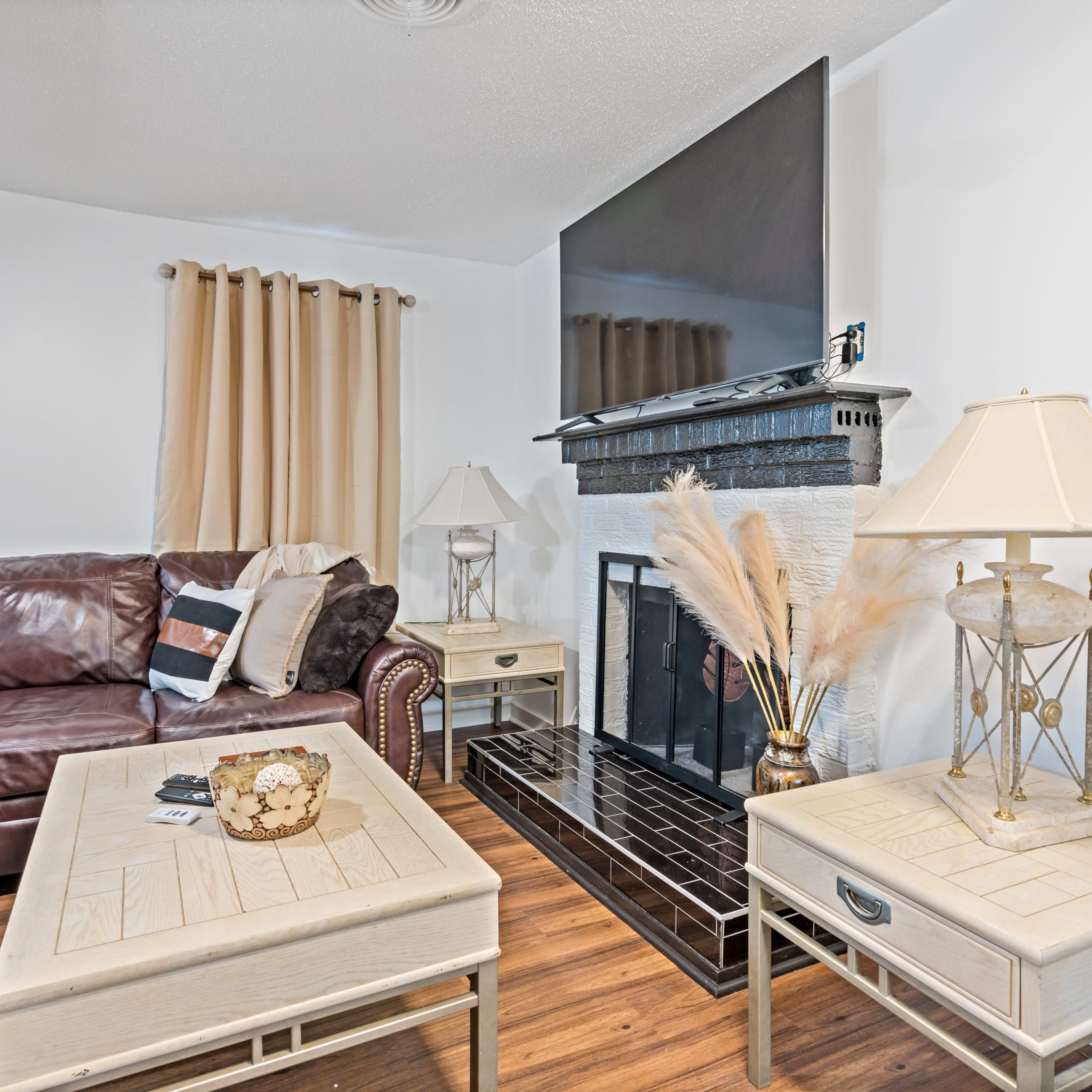 A living room featuring a brown leather sofa, a coffee table with decorative items, and a fireplace surrounded by a black mantel, all set against a backdrop of beige curtains and wood flooring.