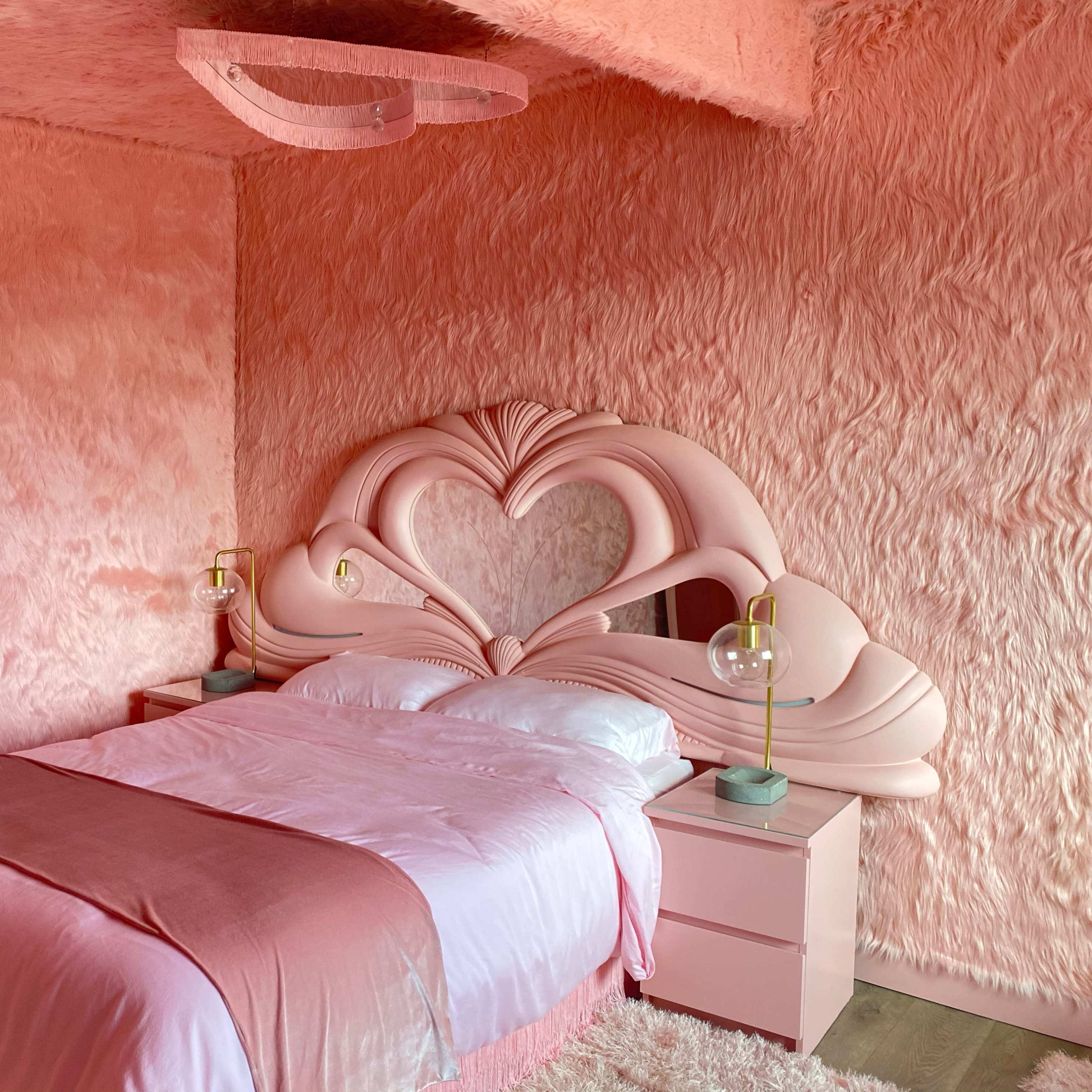 A bedroom with pink, textured walls, a heart-shaped headboard, and a light pink bedspread.