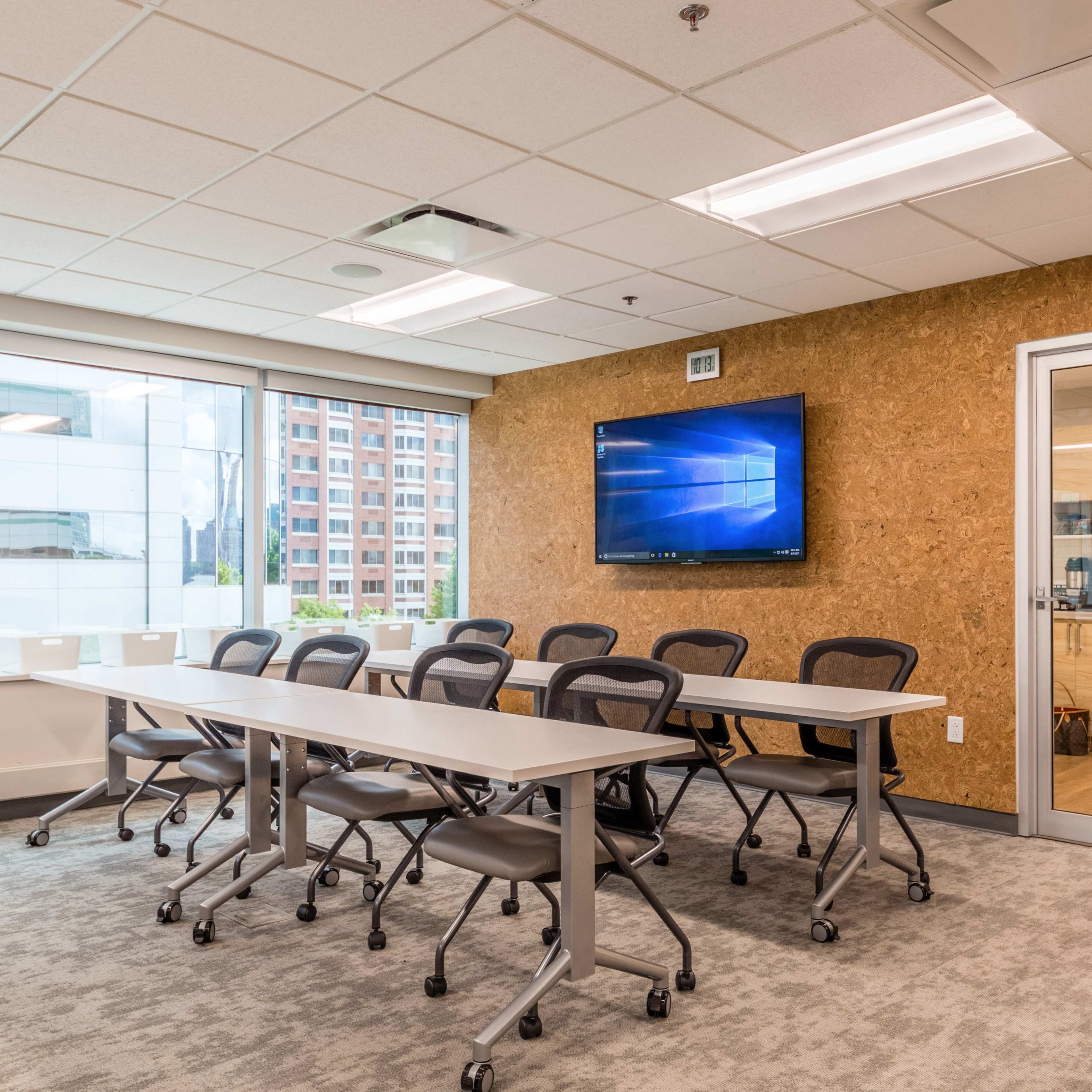 A modern conference room features a long table with rolling chairs, a large screen mounted on the wall, and windows offering a view of a cityscape.