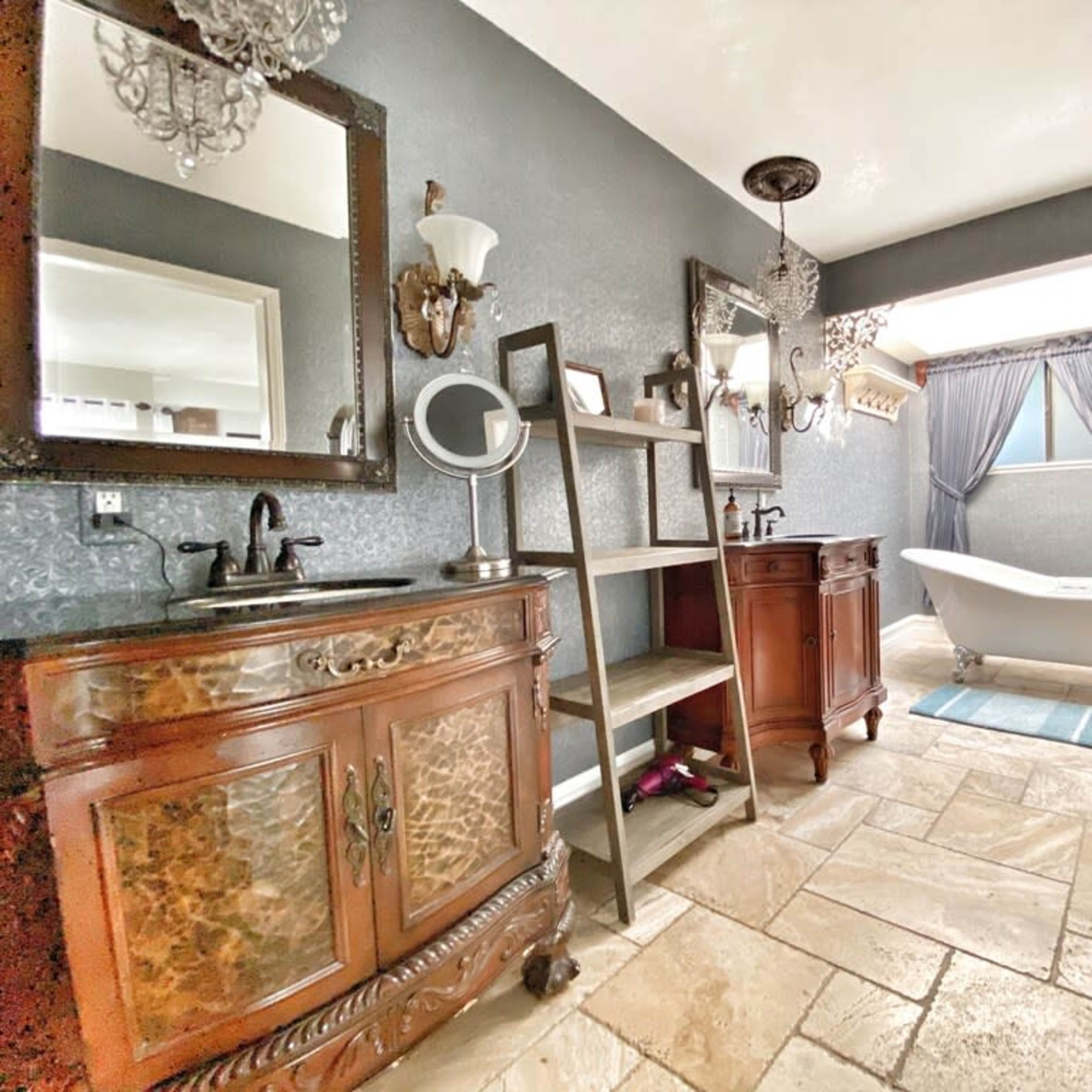 The bathroom features a large freestanding tub, a wooden vanity with an elaborate mirror, and a shelf unit against a textured gray wall.