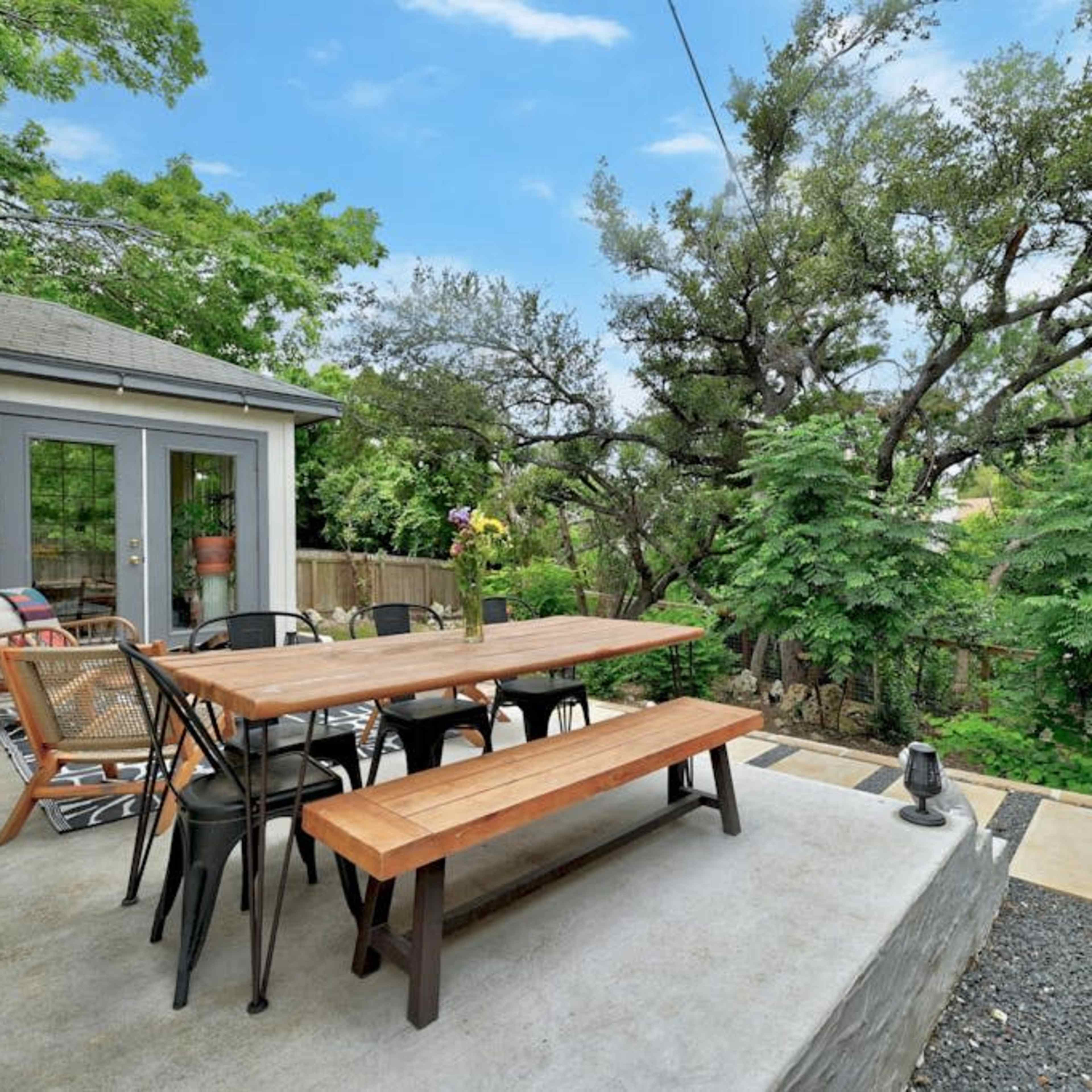 A patio area with a wooden dining table and chairs surrounded by lush greenery and trees.
