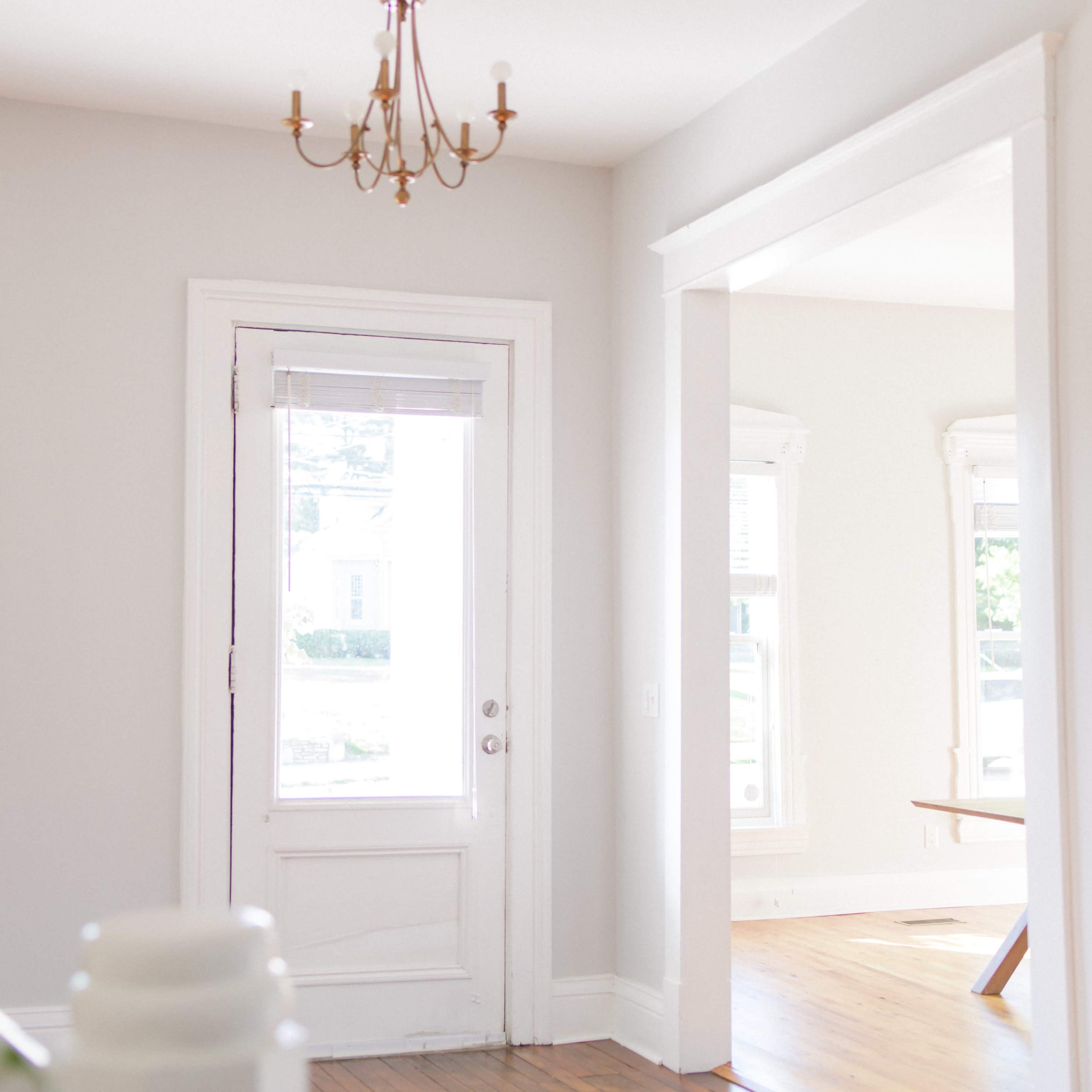 A bright hallway features a front door, a chandelier, and large windows with plants nearby.