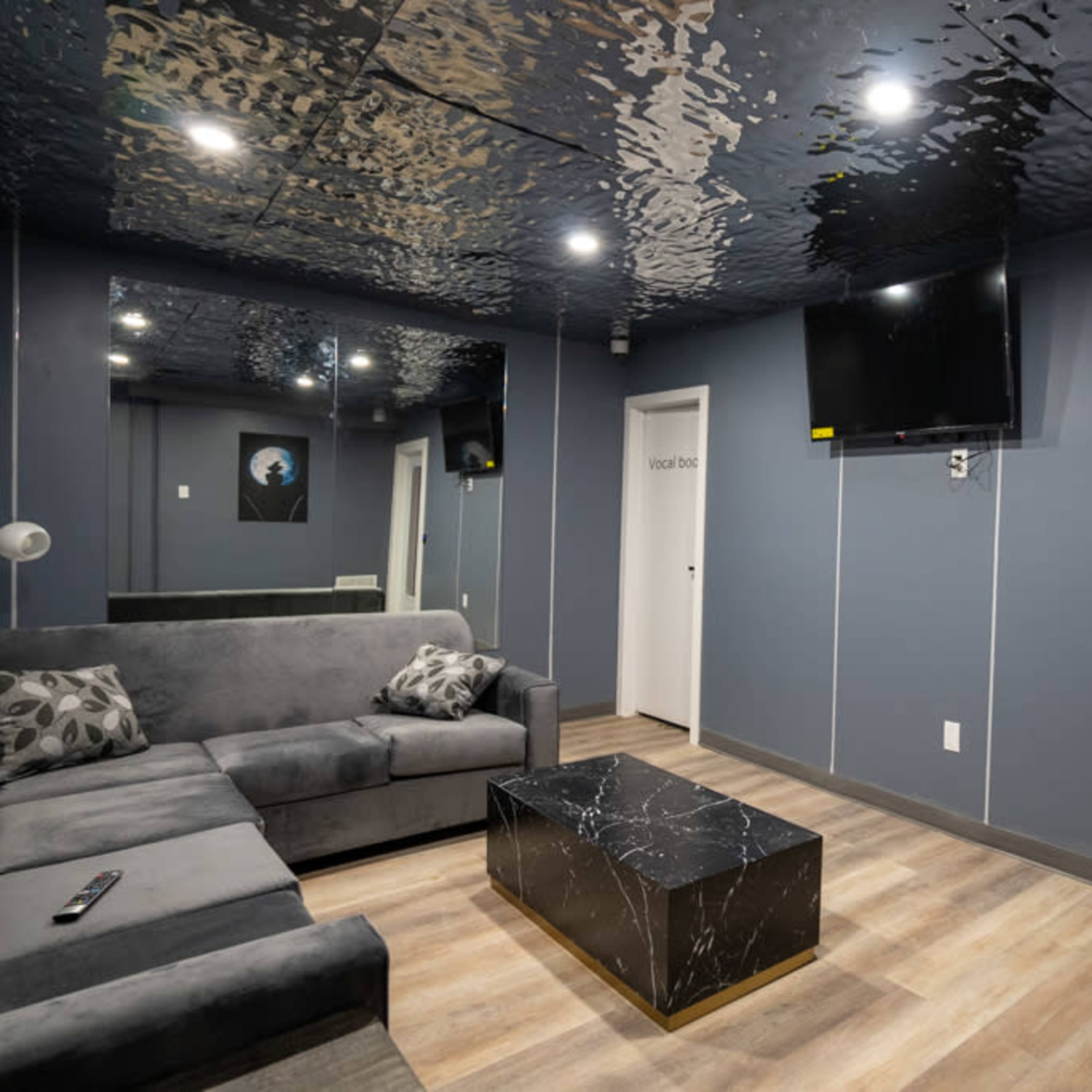 A modern lounge area features a gray sectional sofa, a black marble coffee table, a flat-screen TV, and a door leading to another room, all under a reflective ceiling.