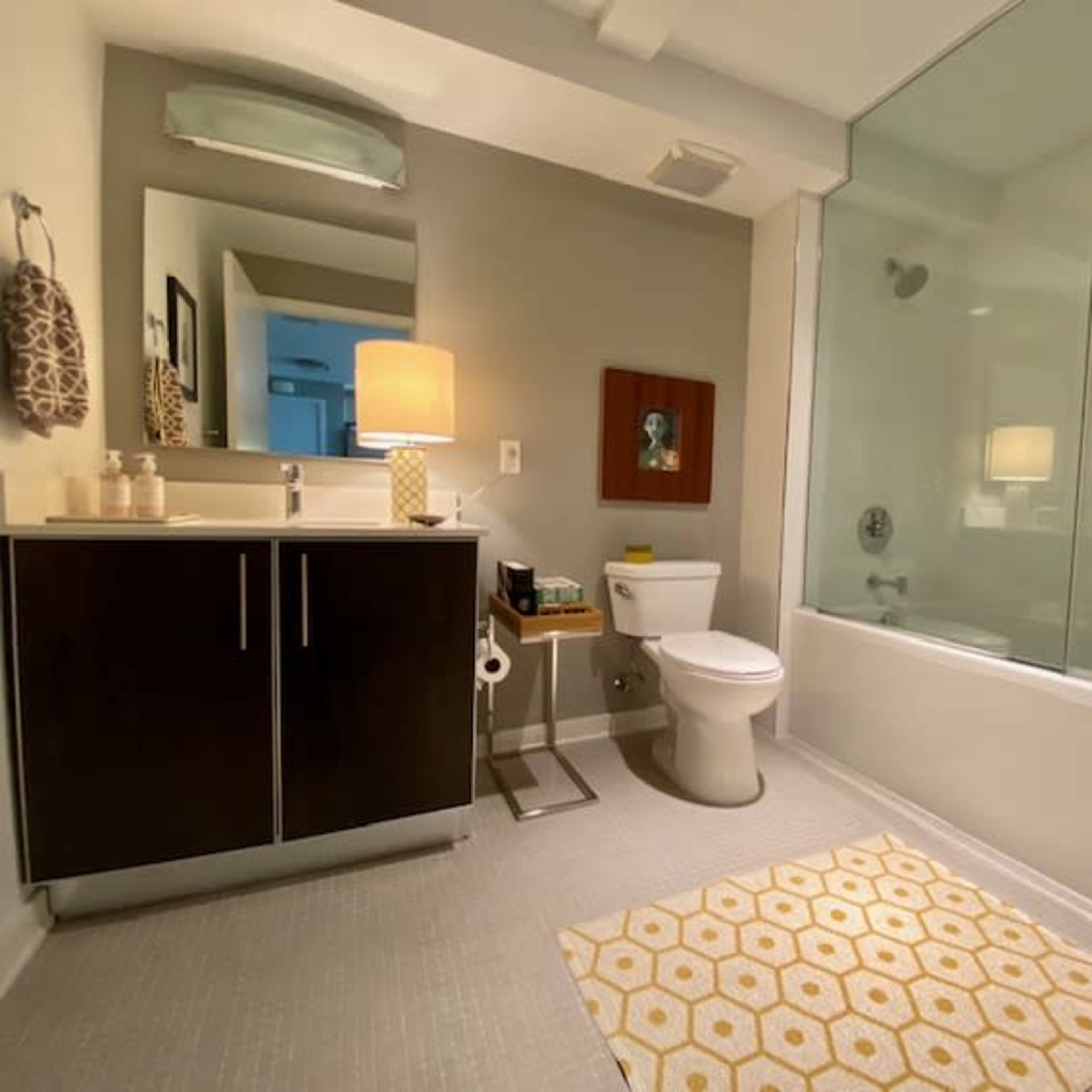 A modern bathroom featuring a glass-enclosed shower, a white toilet, a dark vanity with a sink, and a geometric rug on the floor.