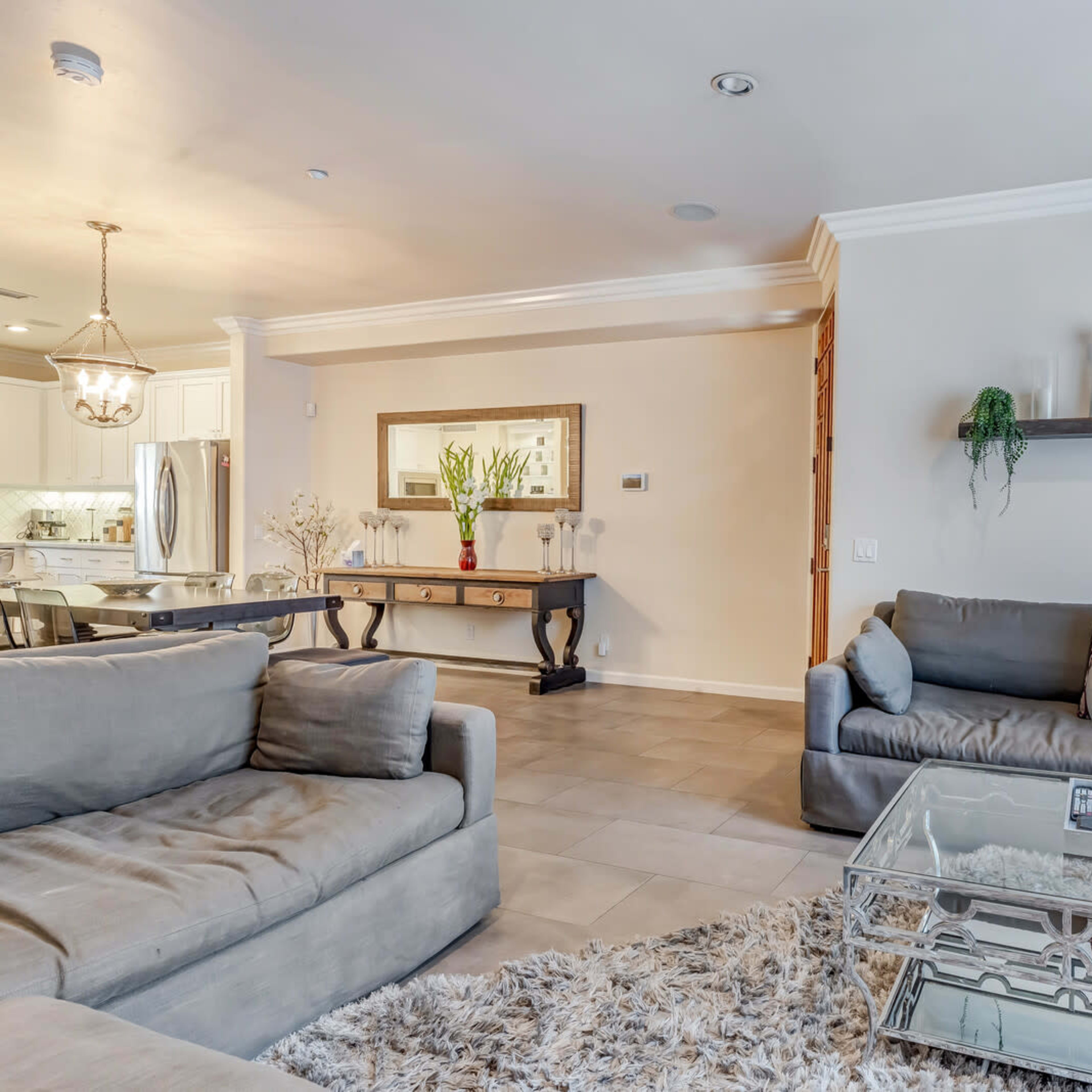 A contemporary living room features two gray sofas, a glass coffee table, and a decorative mirror on the wall, with adjacent kitchen visible in the background.