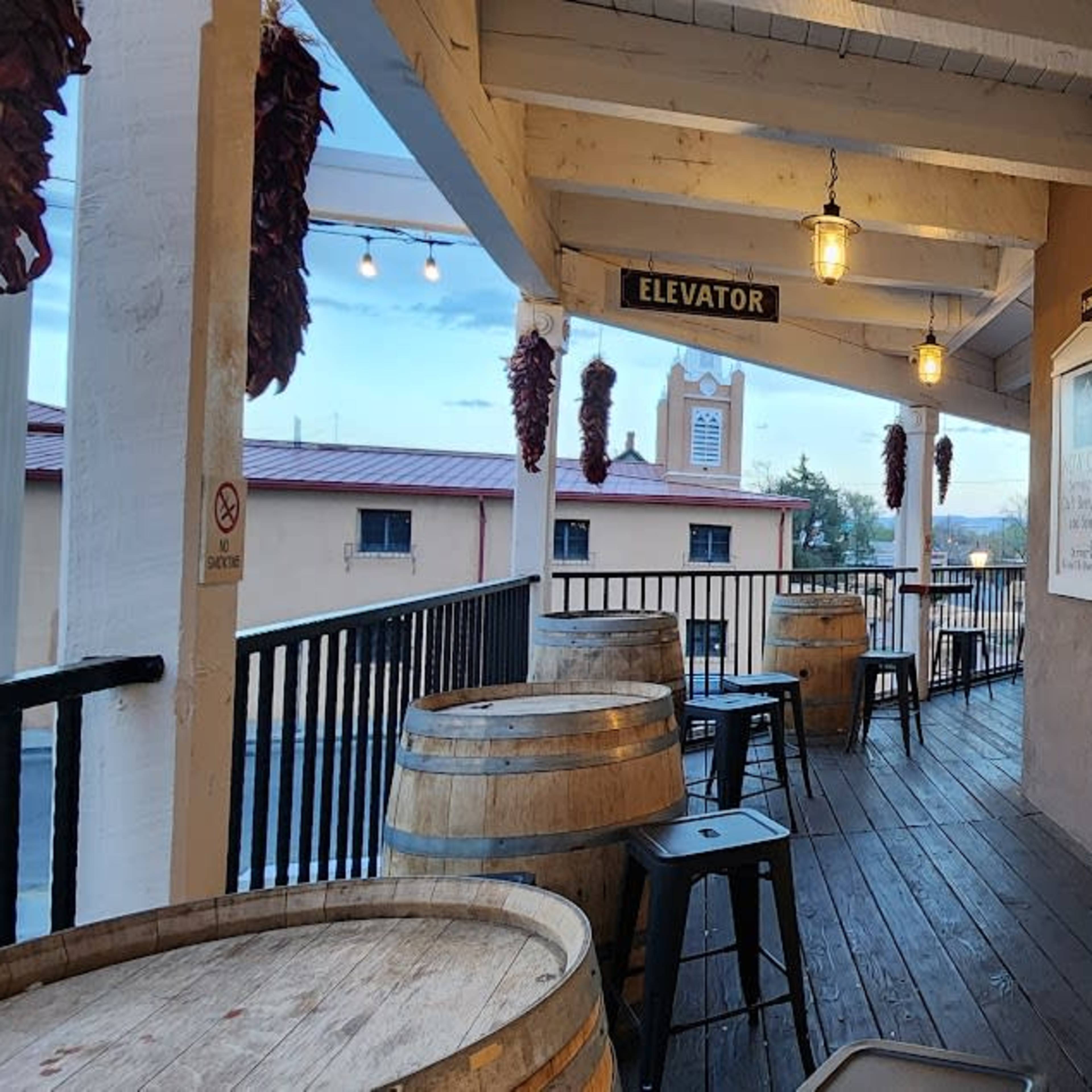 A balcony area with wooden flooring, barrel tables, and a window overlooking a restaurant interior, with a view of buildings in the background.