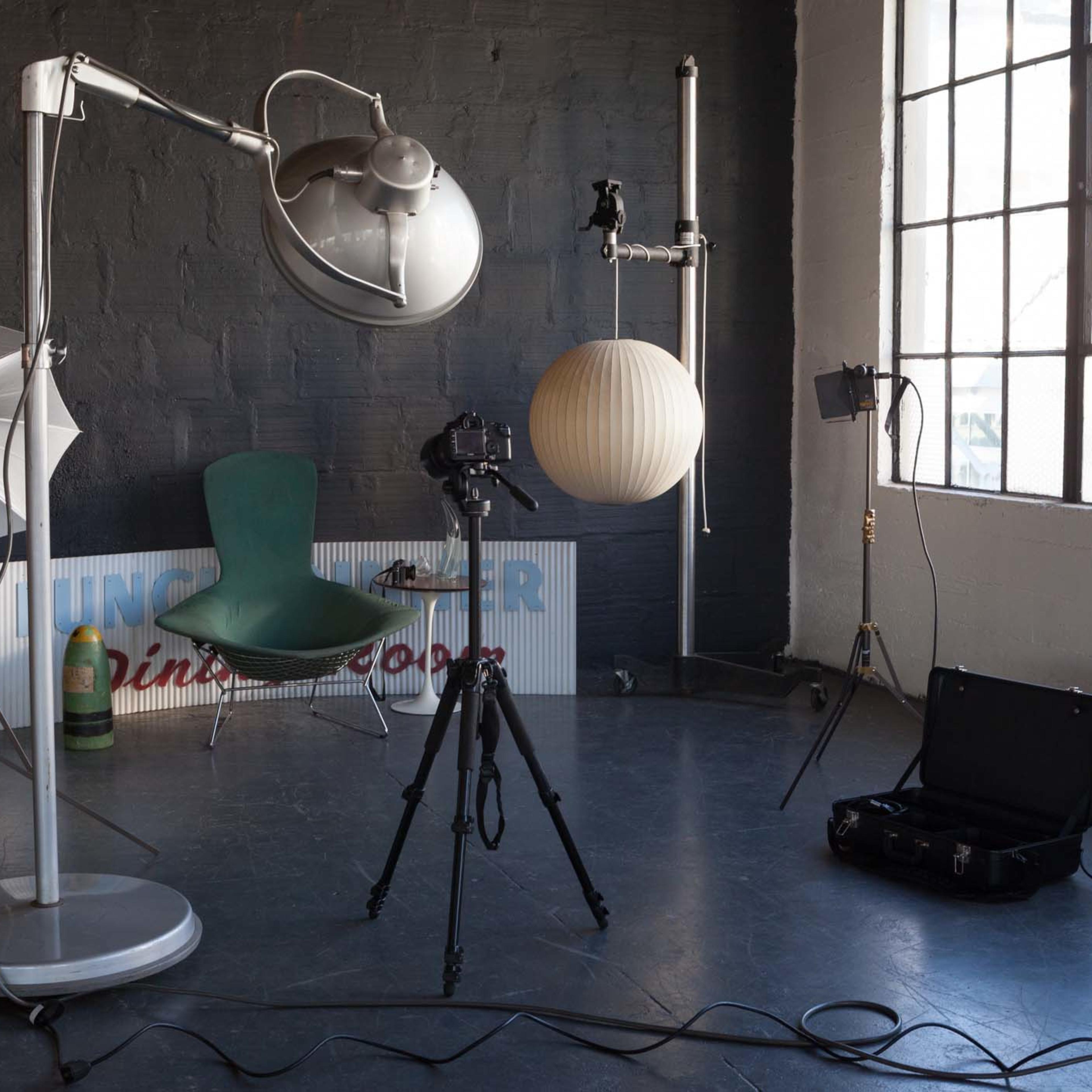 A photography studio setup with a camera on a tripod, a large light umbrella, and a round paper lamp hanging from the ceiling.