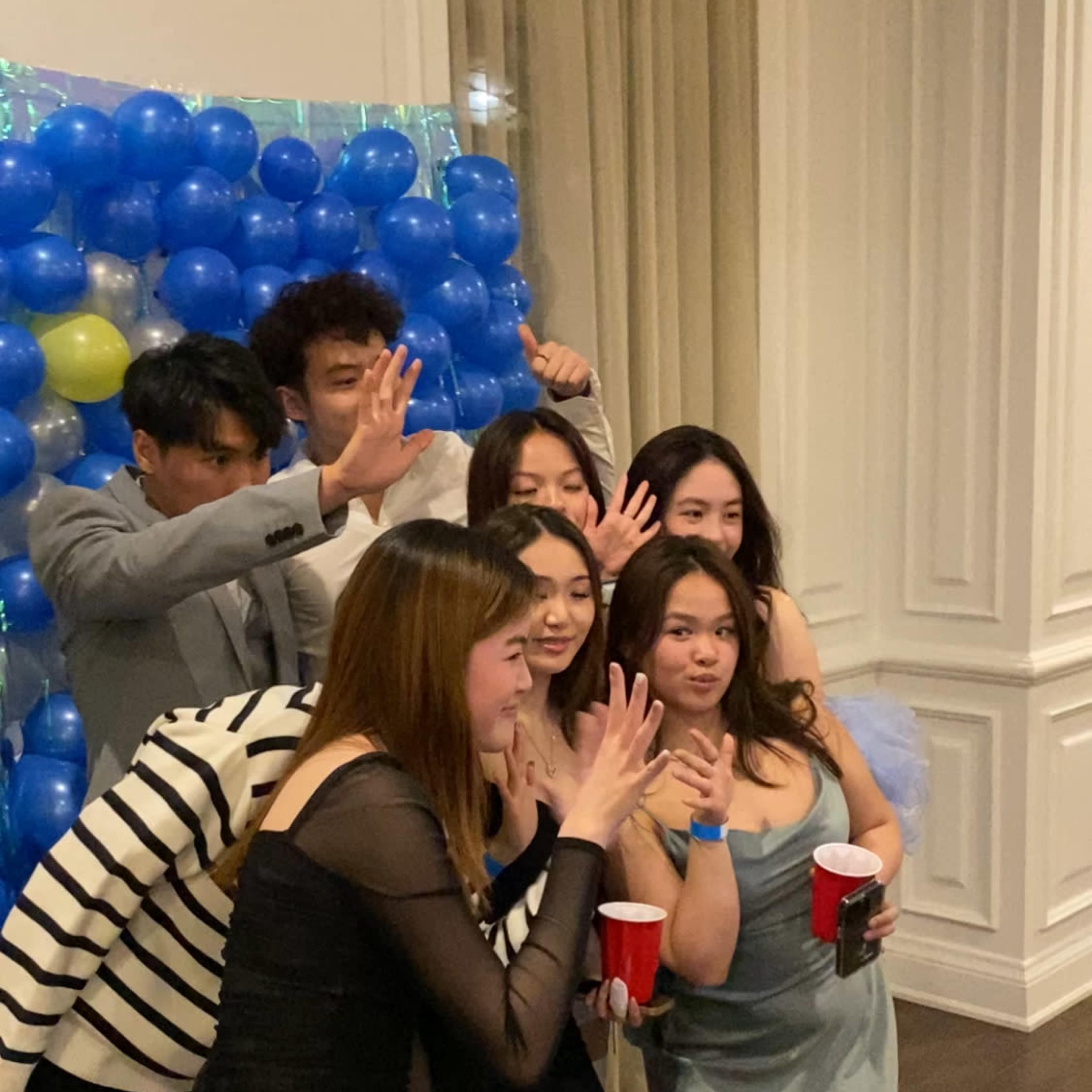 A group of six young adults poses together in front of a colorful balloon display at a social gathering.