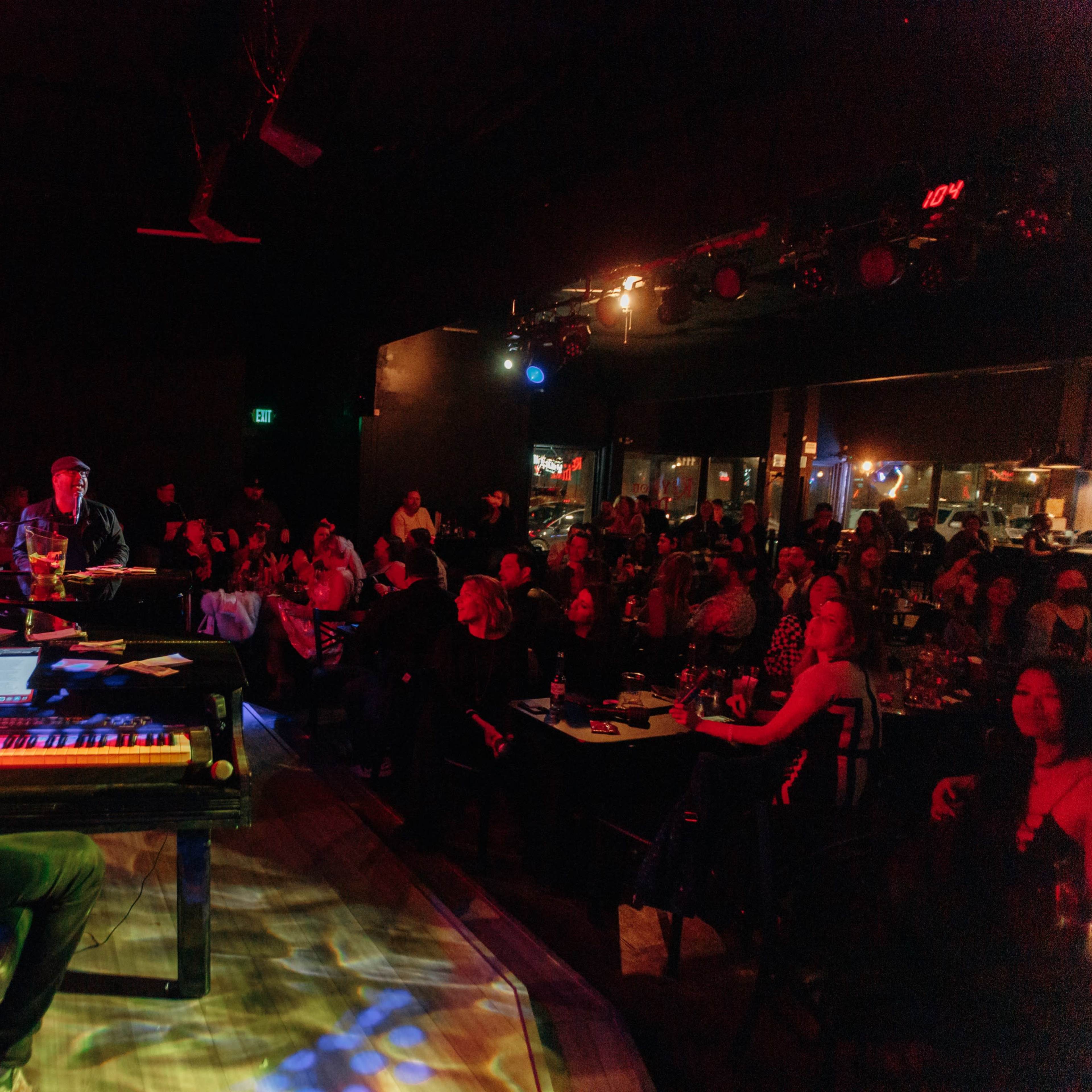 A musician plays a keyboard on stage while an audience sits at tables, attentively watching the performance in a dimly lit venue.