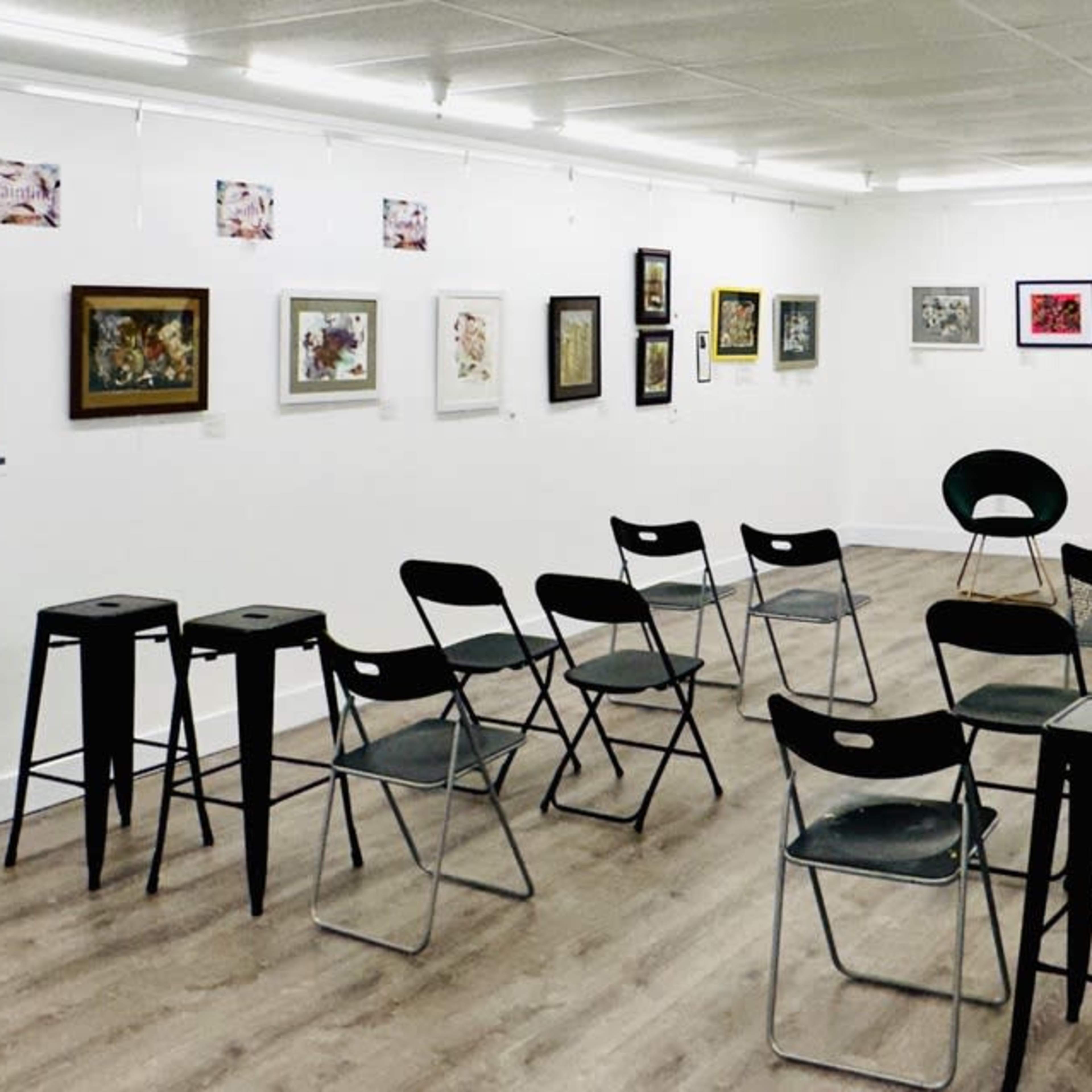 An art gallery with several framed artworks displayed on the walls and rows of black folding chairs set up on the floor.