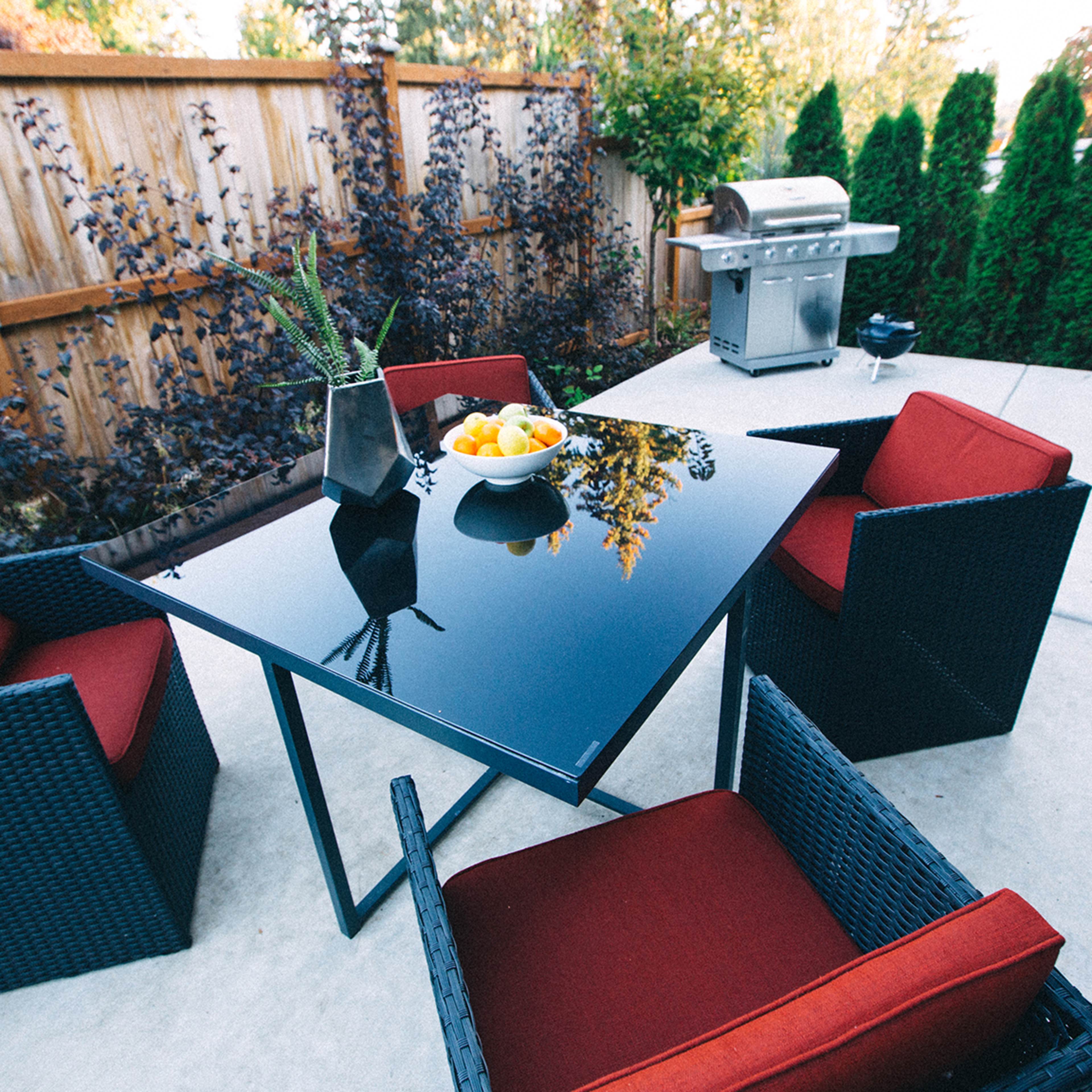 A modern outdoor dining area features a black table surrounded by four red chairs, with a bowl of fruit at the center and a grill in the background.