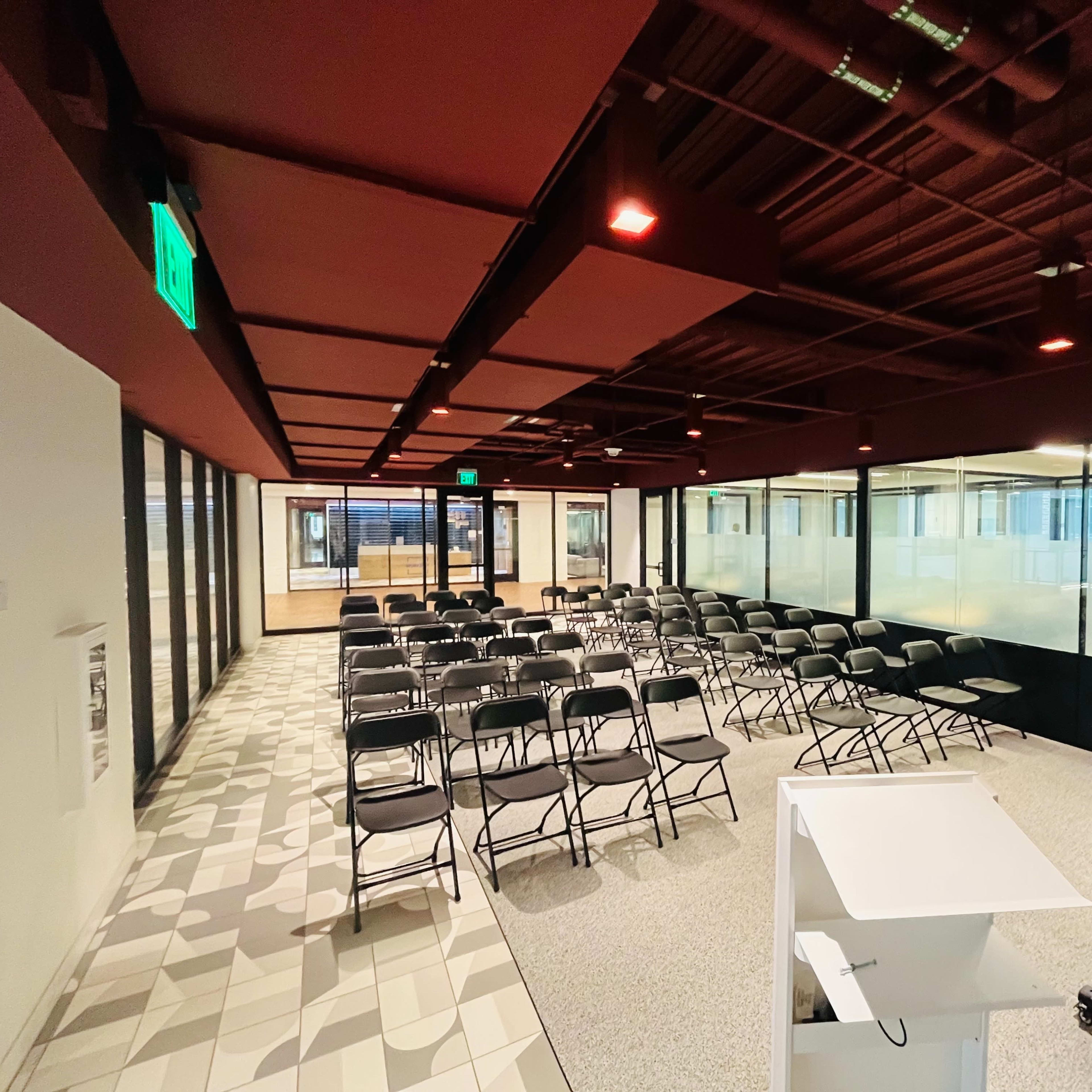 A spacious meeting room with rows of black chairs arranged on a patterned floor, surrounded by glass walls and equipped with a presentation podium.