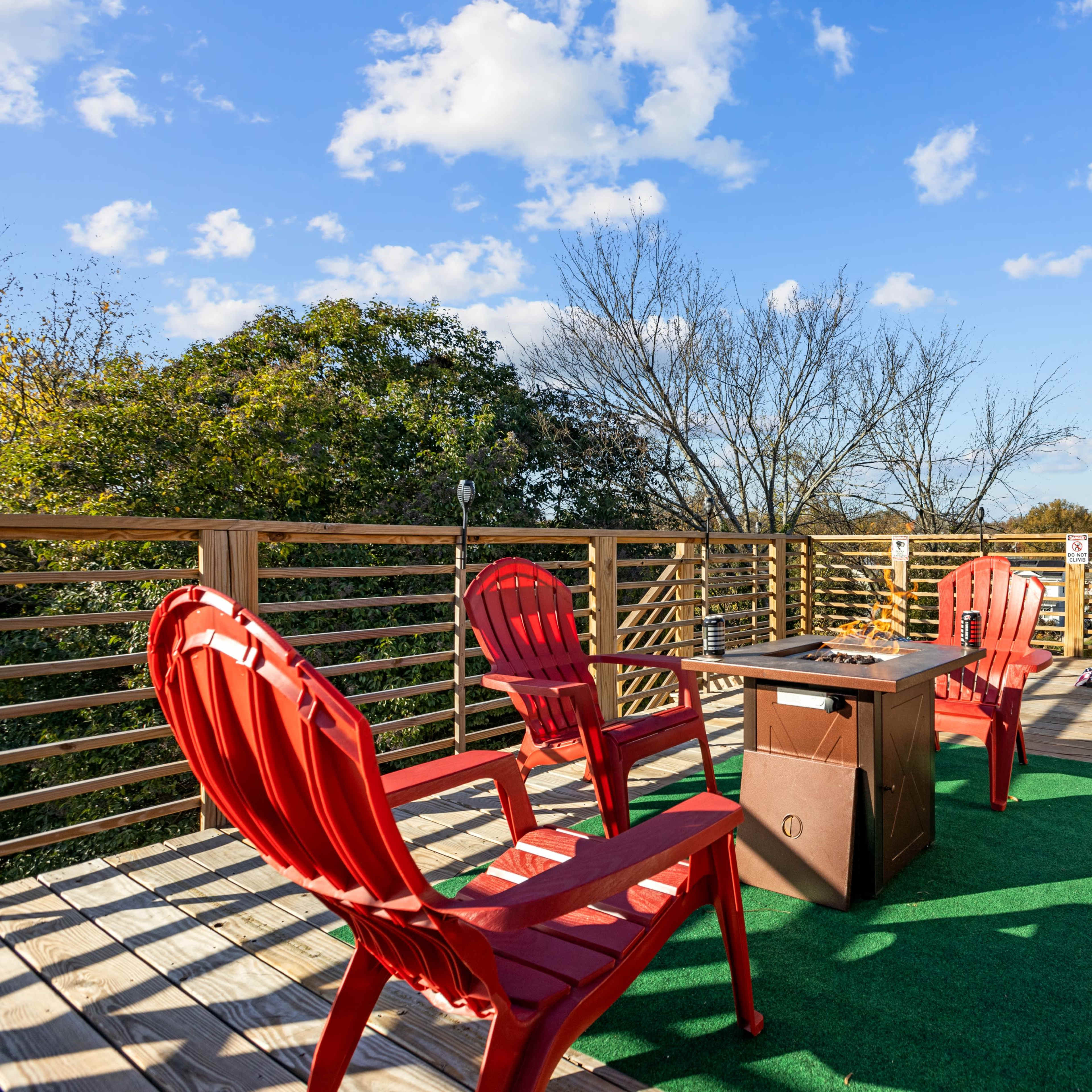 A deck features four red chairs arranged around a central table, with trees and a blue sky in the background.