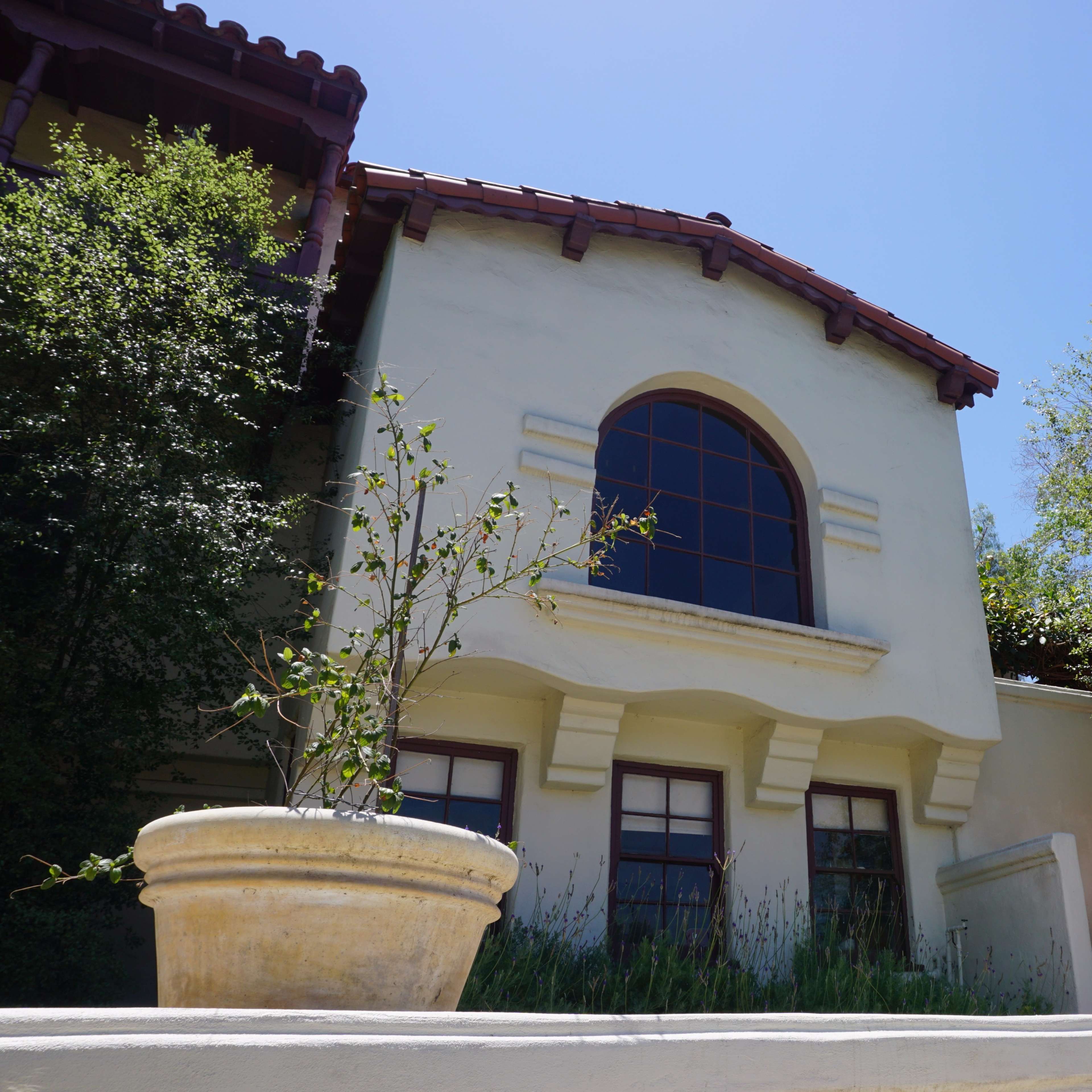A beige house with a curved window and a terracotta roof is surrounded by greenery and features a large planter at its base.