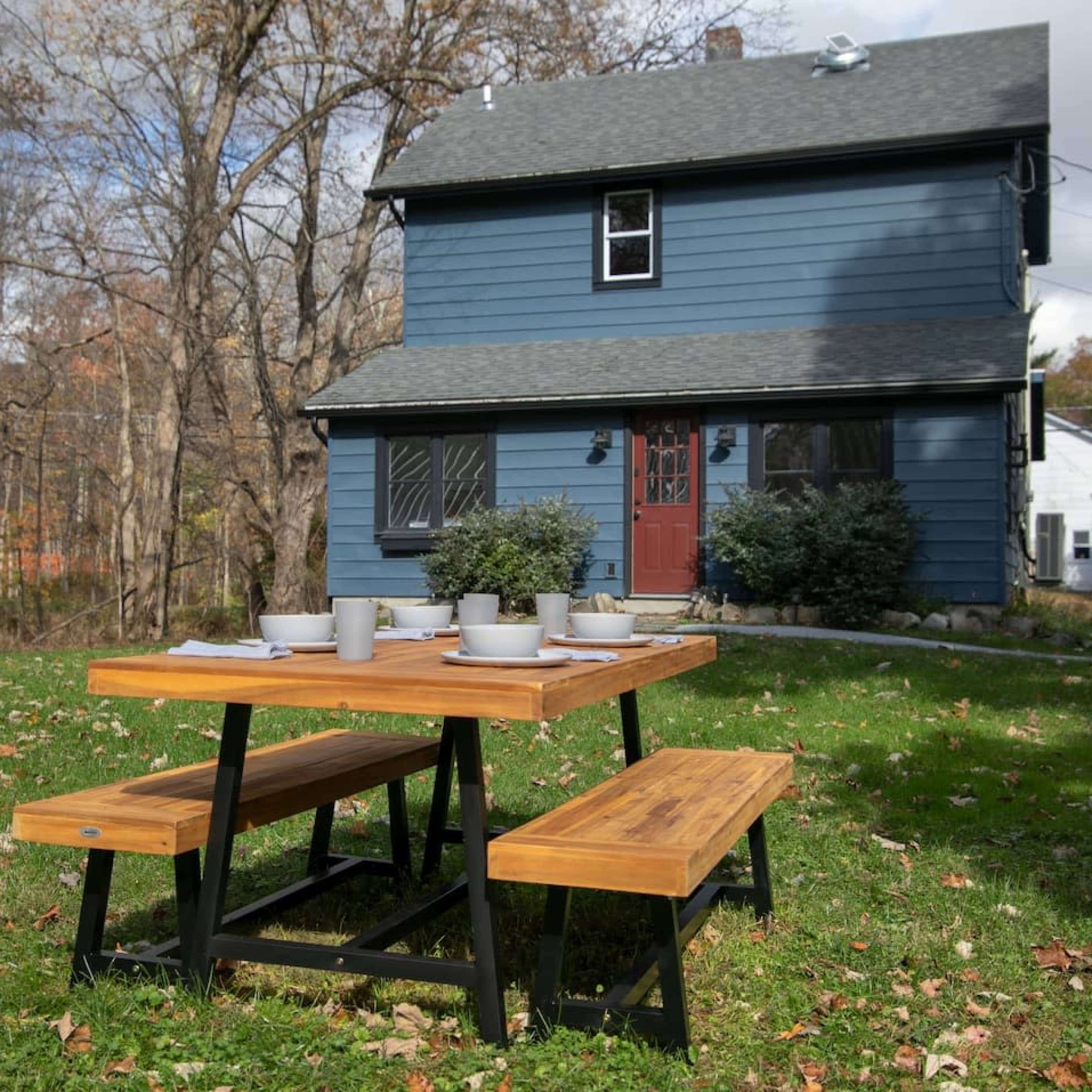 A wooden picnic table with benches is set up on the grass in front of a blue house surrounded by trees.