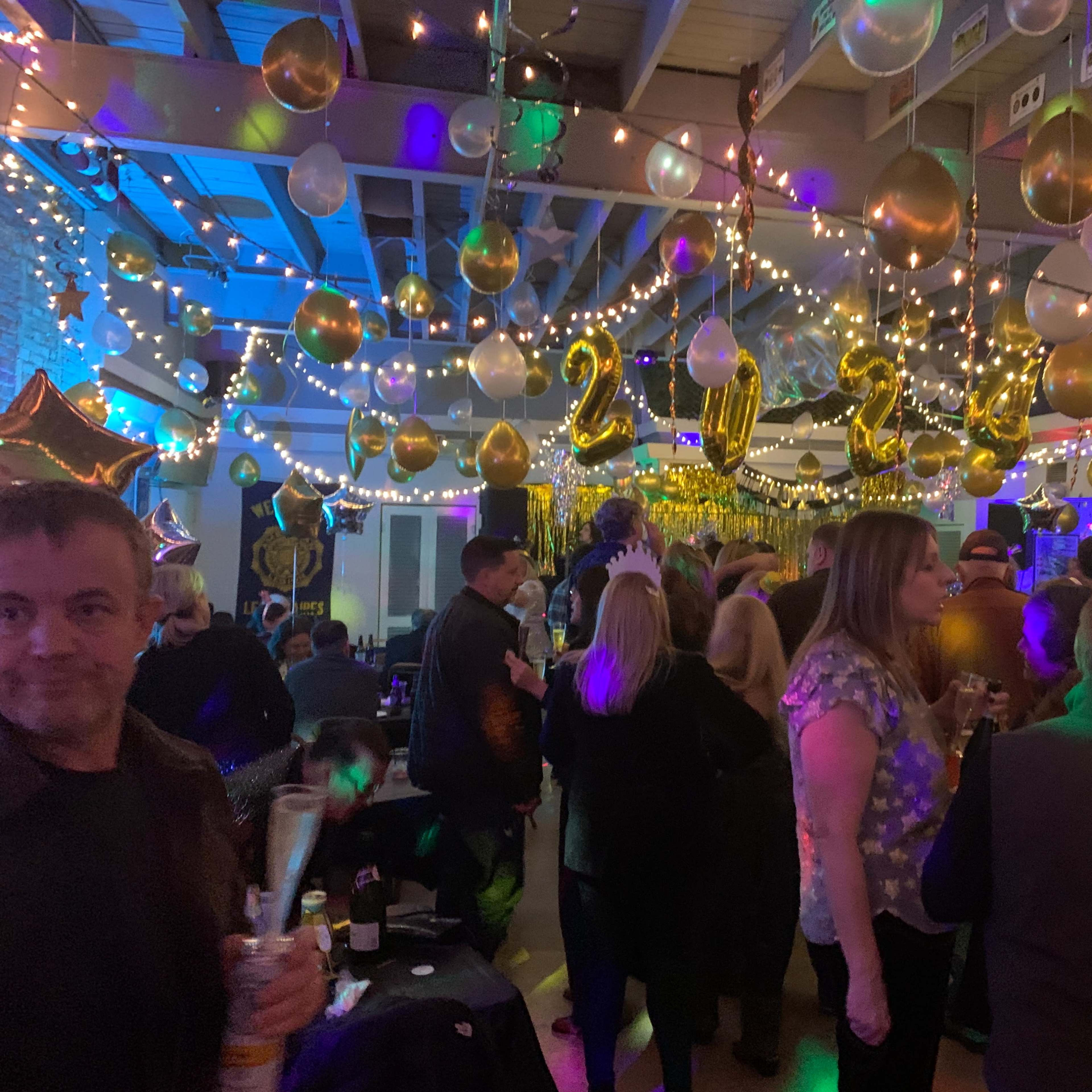 A crowded indoor party features balloons and string lights, with guests celebrating the arrival of the year 2023.
