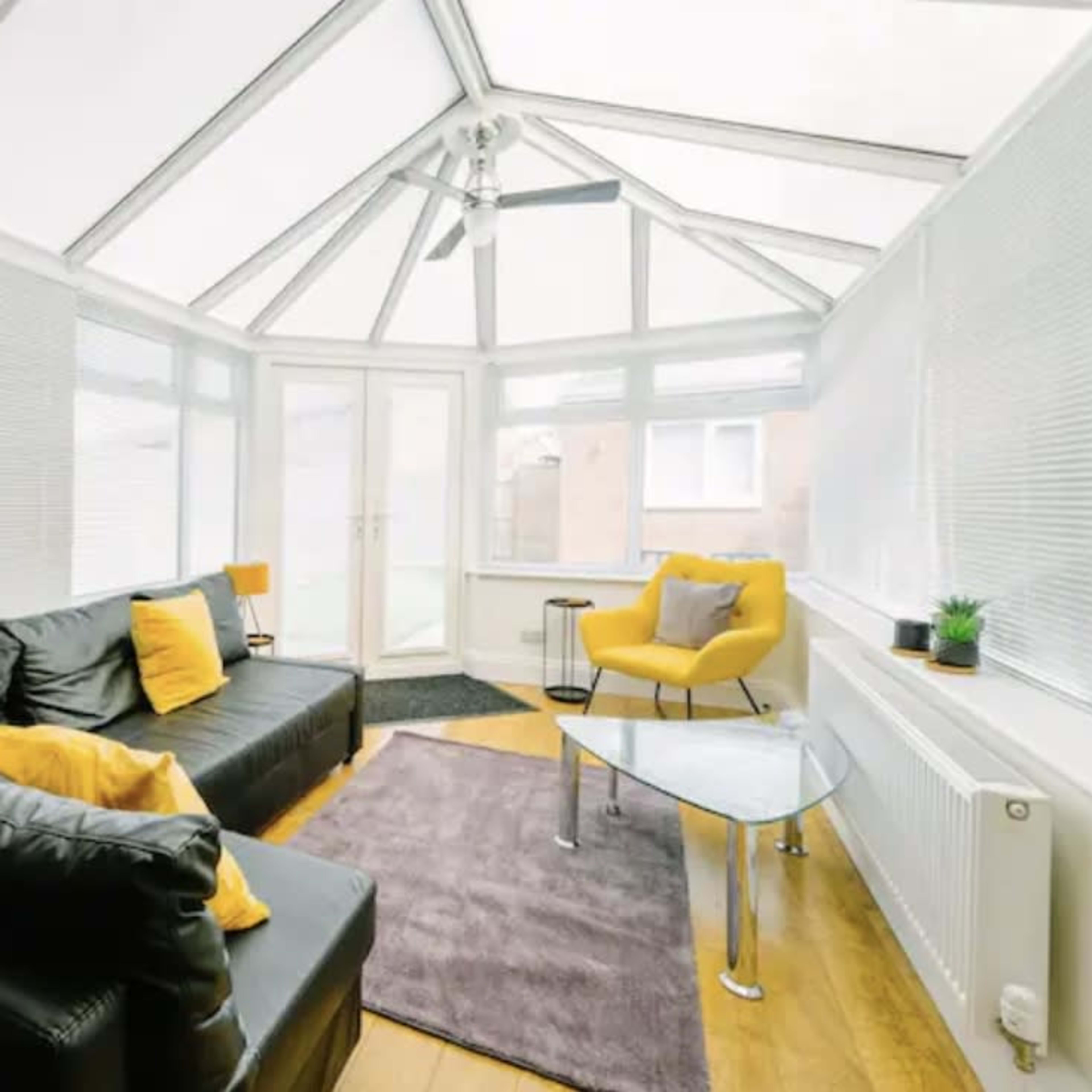 A bright conservatory with a black leather sofa, a yellow accent chair, and a glass coffee table on a rug.