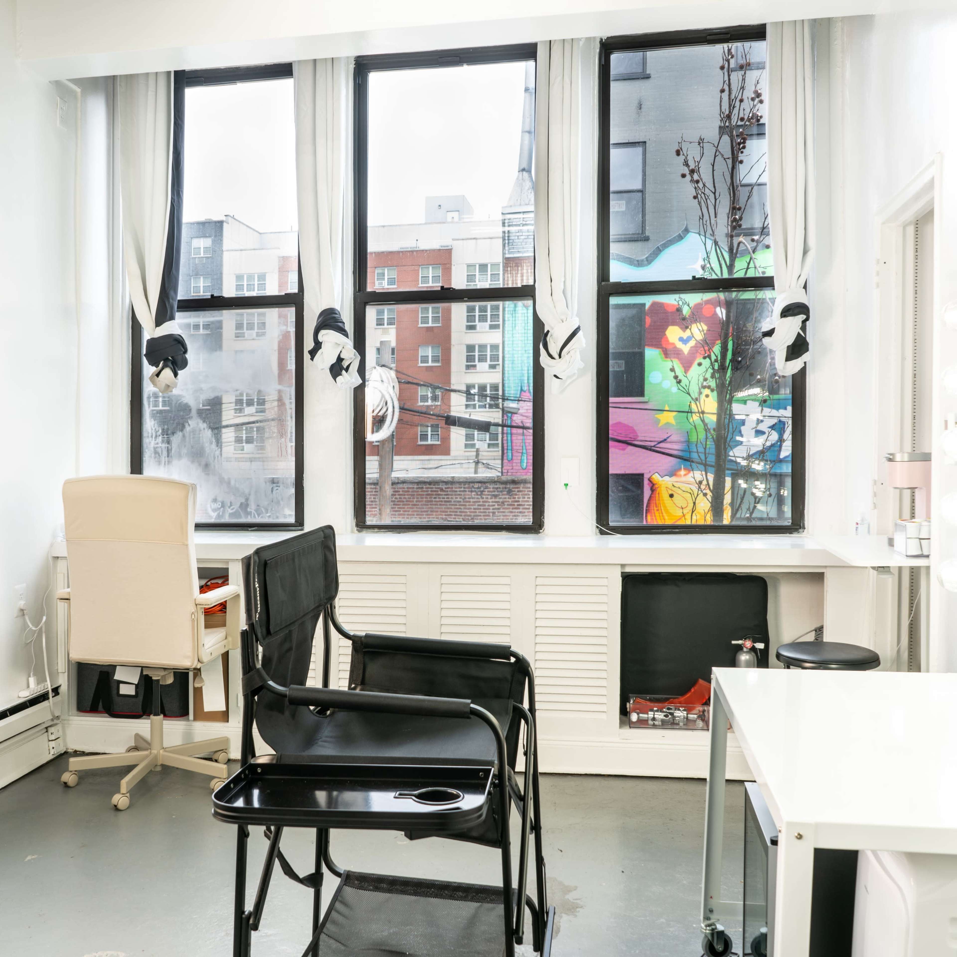 A bright, minimalistic room with large windows displaying urban scenery, featuring a few chairs and a vanity setup.