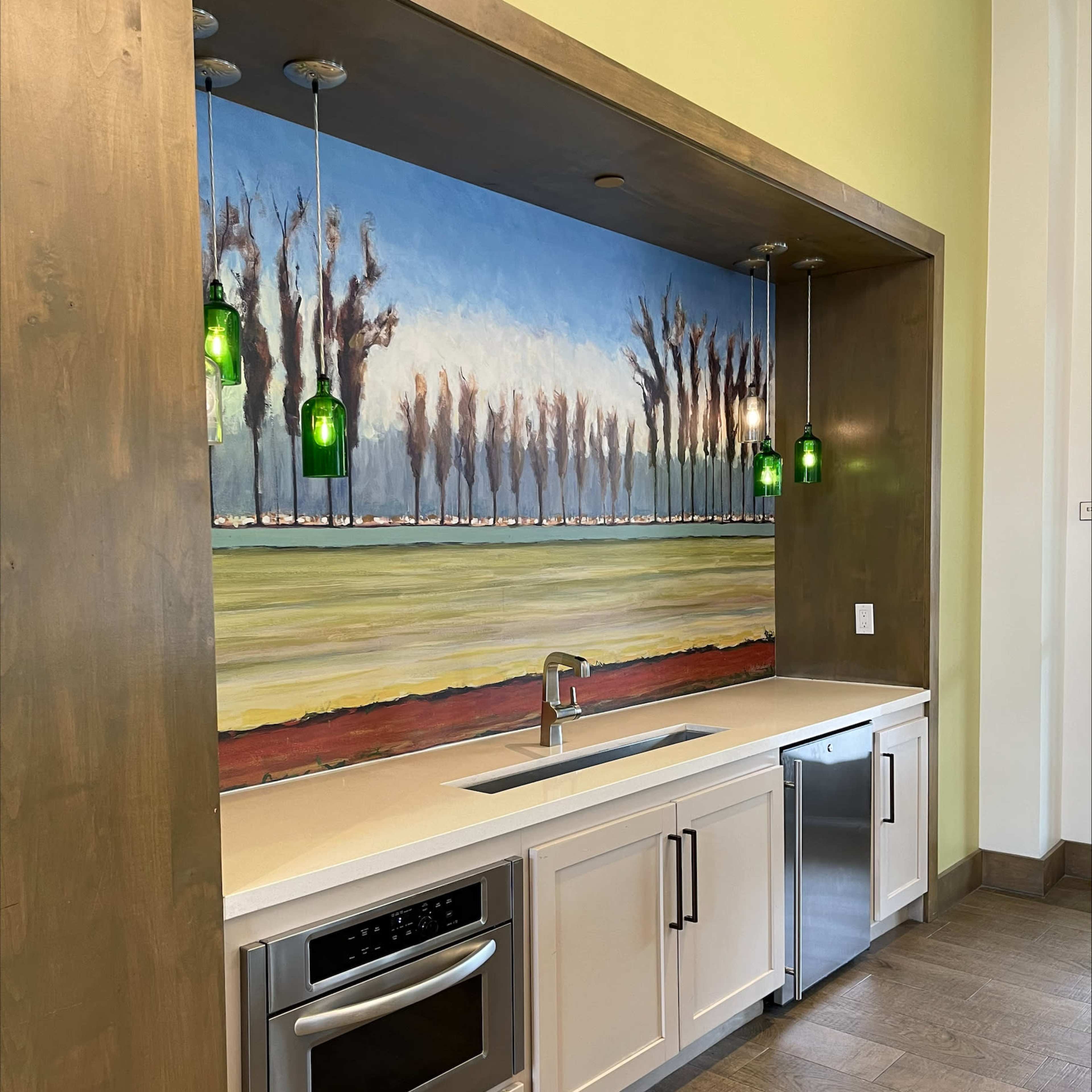 A modern kitchen with a counter, a sink, a microwave, and a refrigerator, featuring a mural of trees and fields above the backdrop.