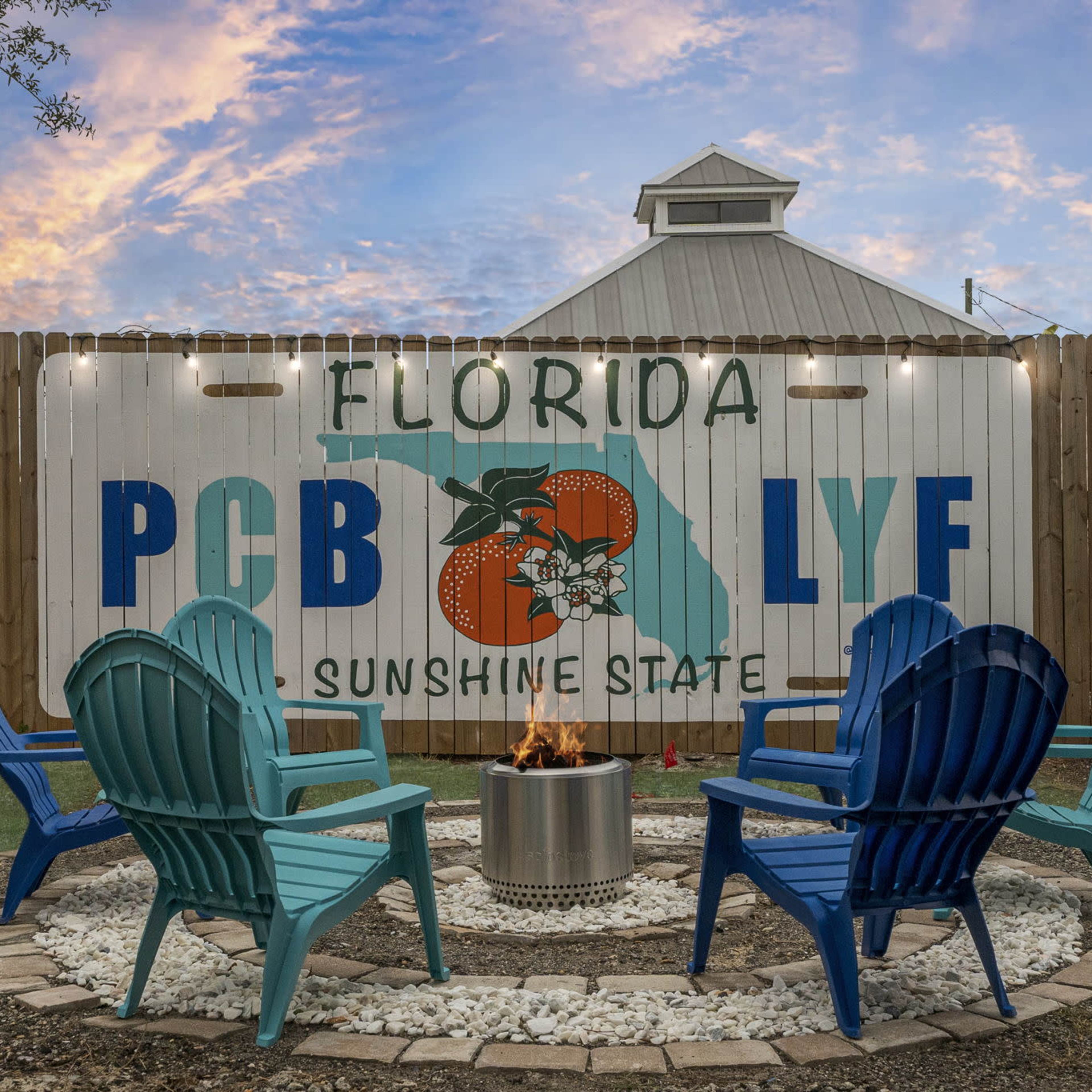 A cozy outdoor fire pit area with blue and green adirondack chairs arranged around a circular stone layout, featuring a mural of the Florida map and the words "Florida PCB LYF Sunshine State" on a wooden fence in the background.
