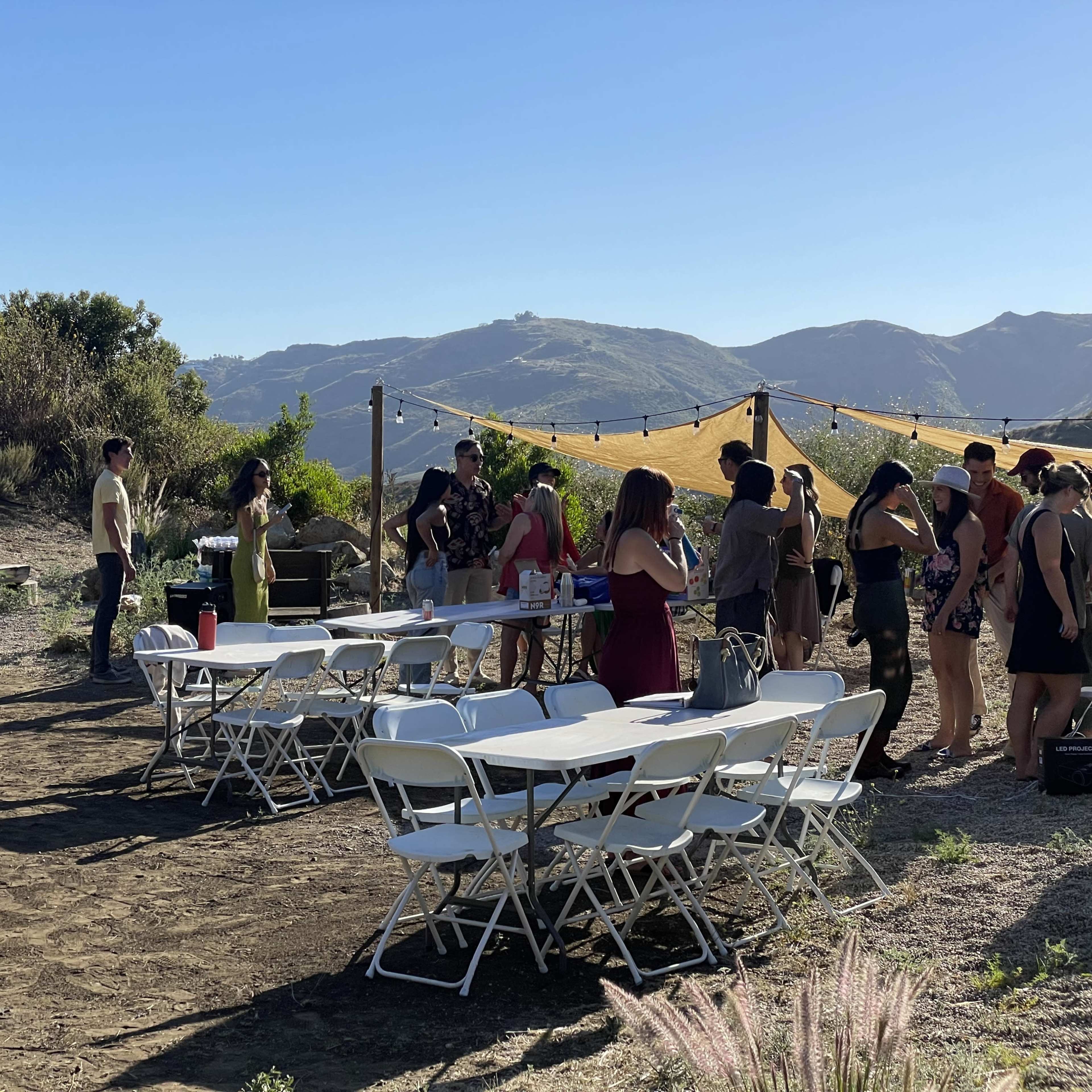 A group of people gathers around tables set up outdoors on a hillside with a view of mountains in the background.