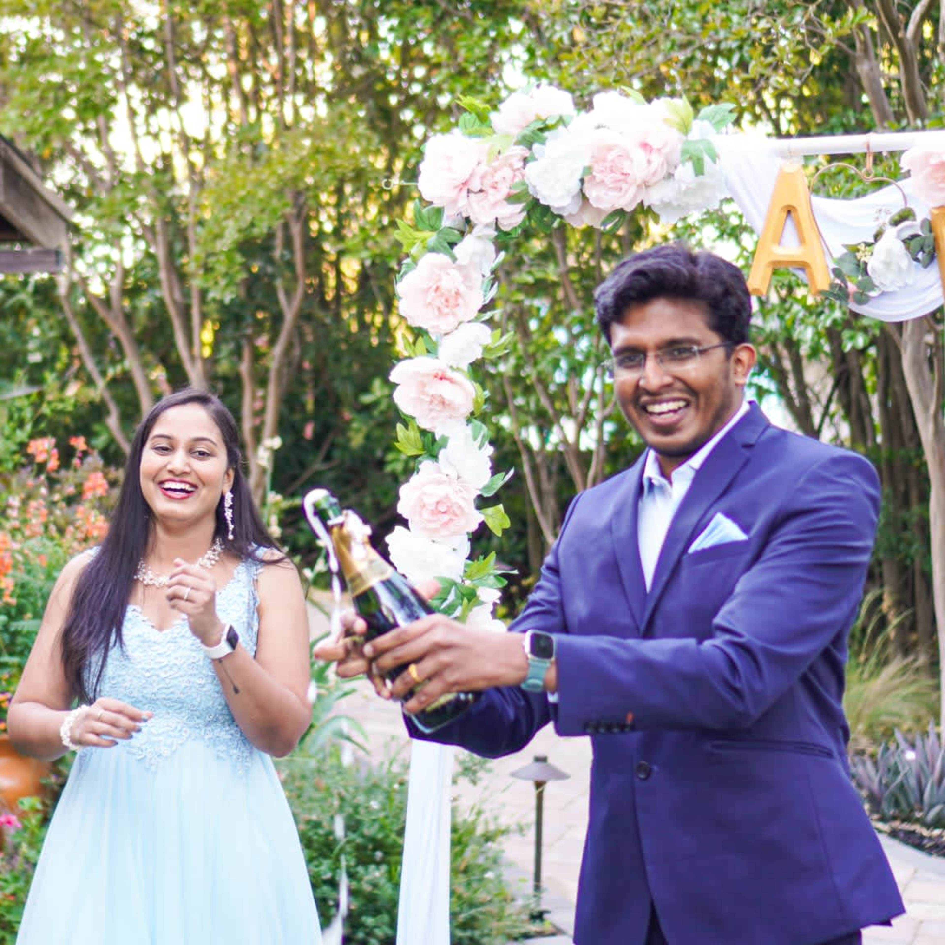 A man in a suit opens a champagne bottle while a woman in a light blue dress smiles beside him, surrounded by floral decorations in a garden setting.